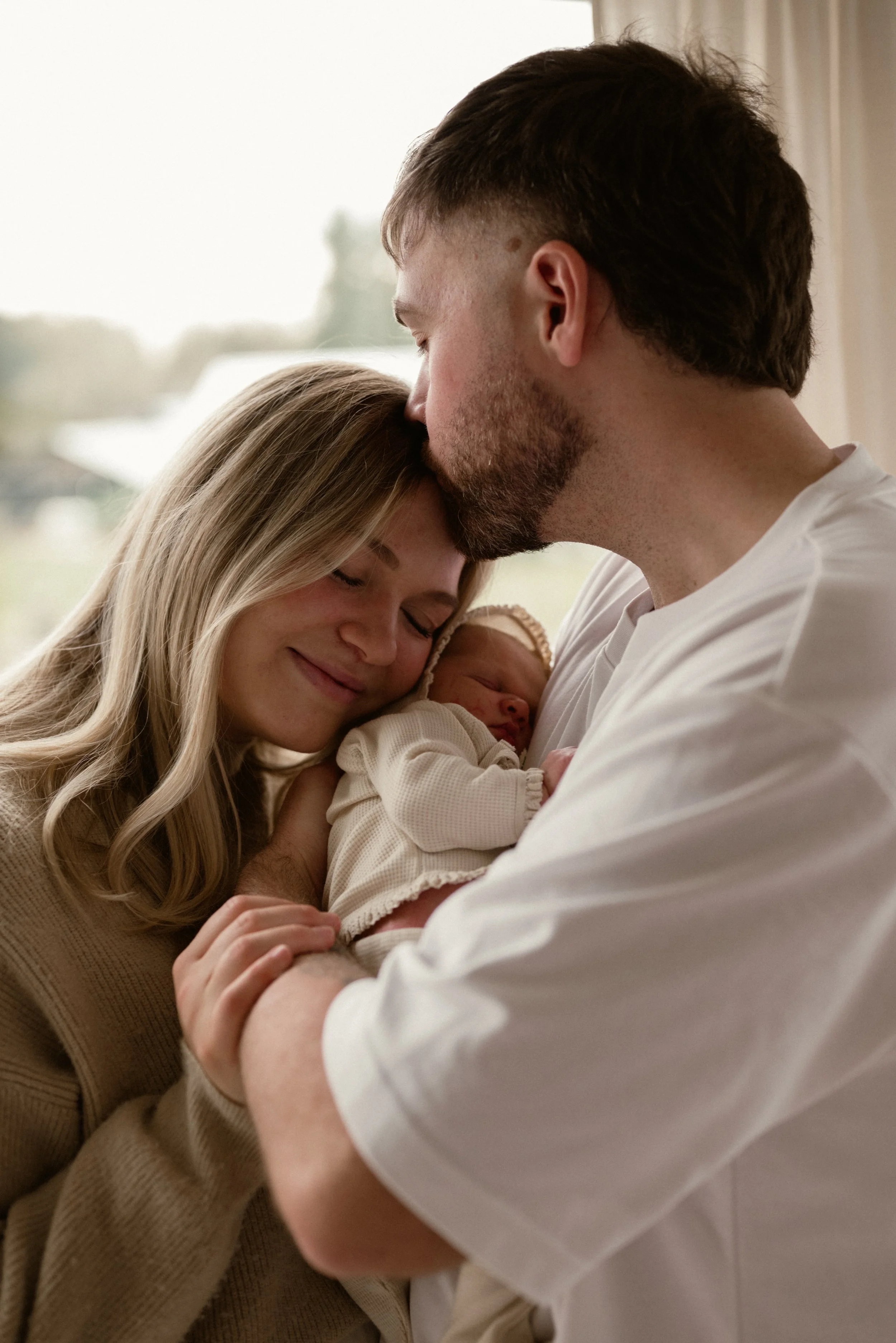 A couple holding a newborn baby with the father kissing the mother on the forehead, in a warmly lit room.