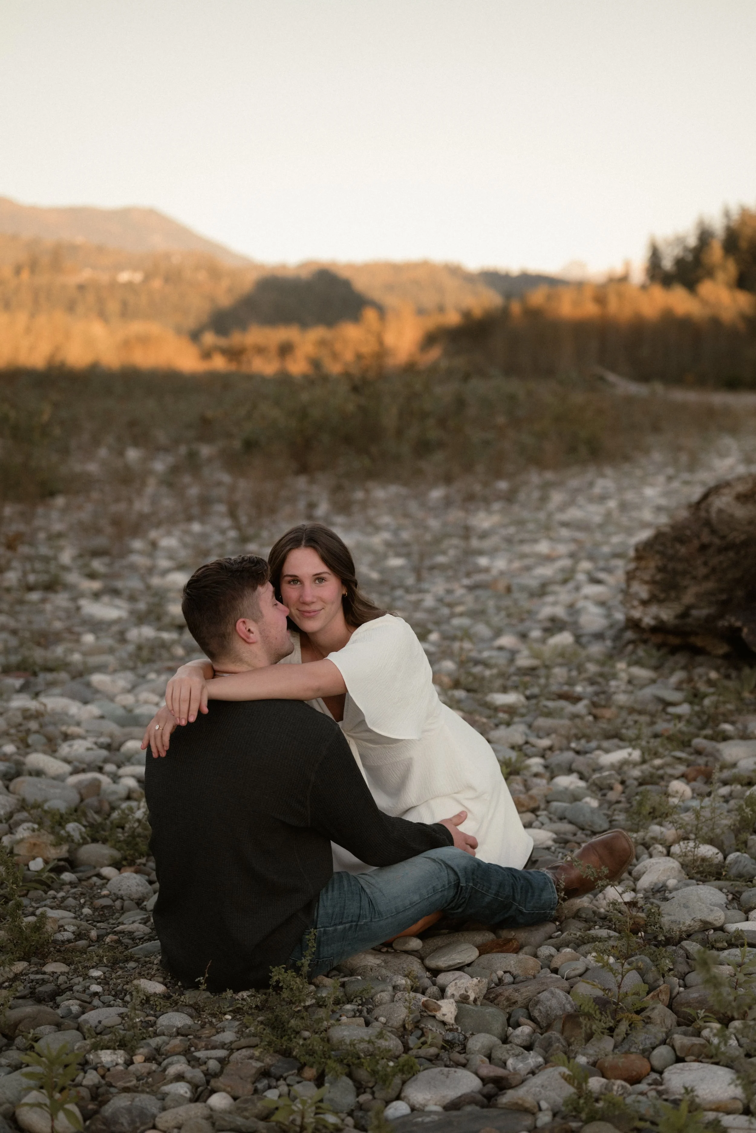 A young couple sitting on a rocky beach at sunset, with mountains in the background. The woman is embracing the man and looking at the camera, while the man has his eyes closed.