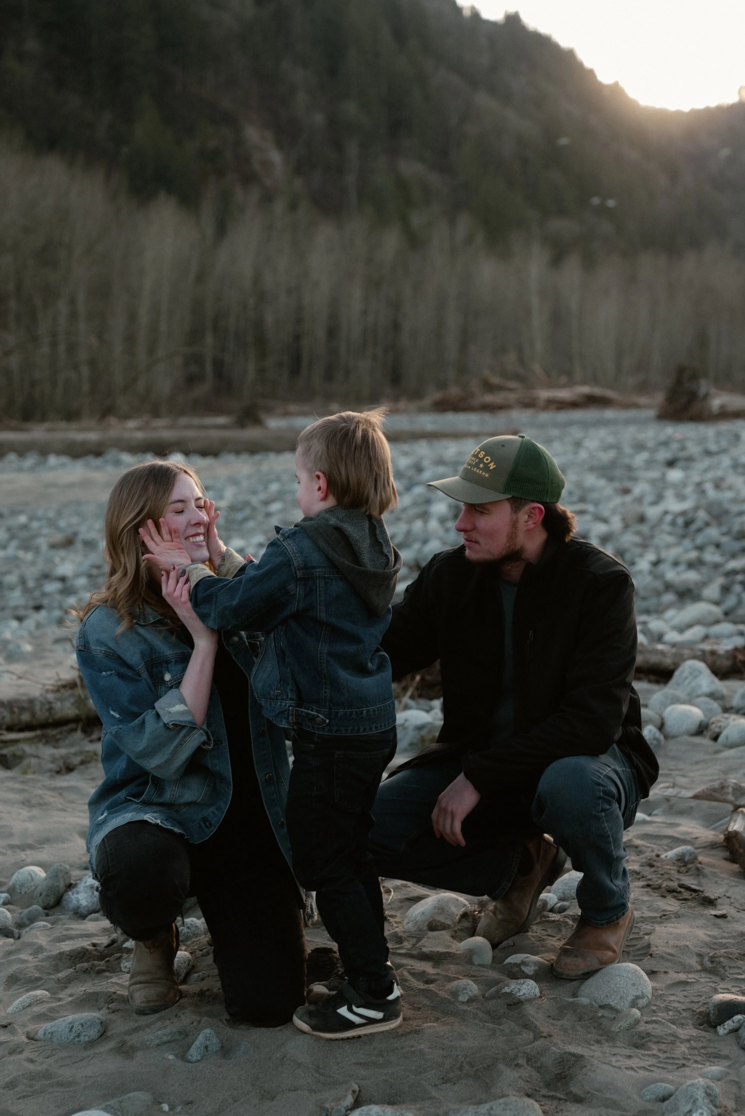 A young family on a rocky riverside at sunset. The mother smiles as her young son touches her face, and the father kneels beside them, wearing a green cap.