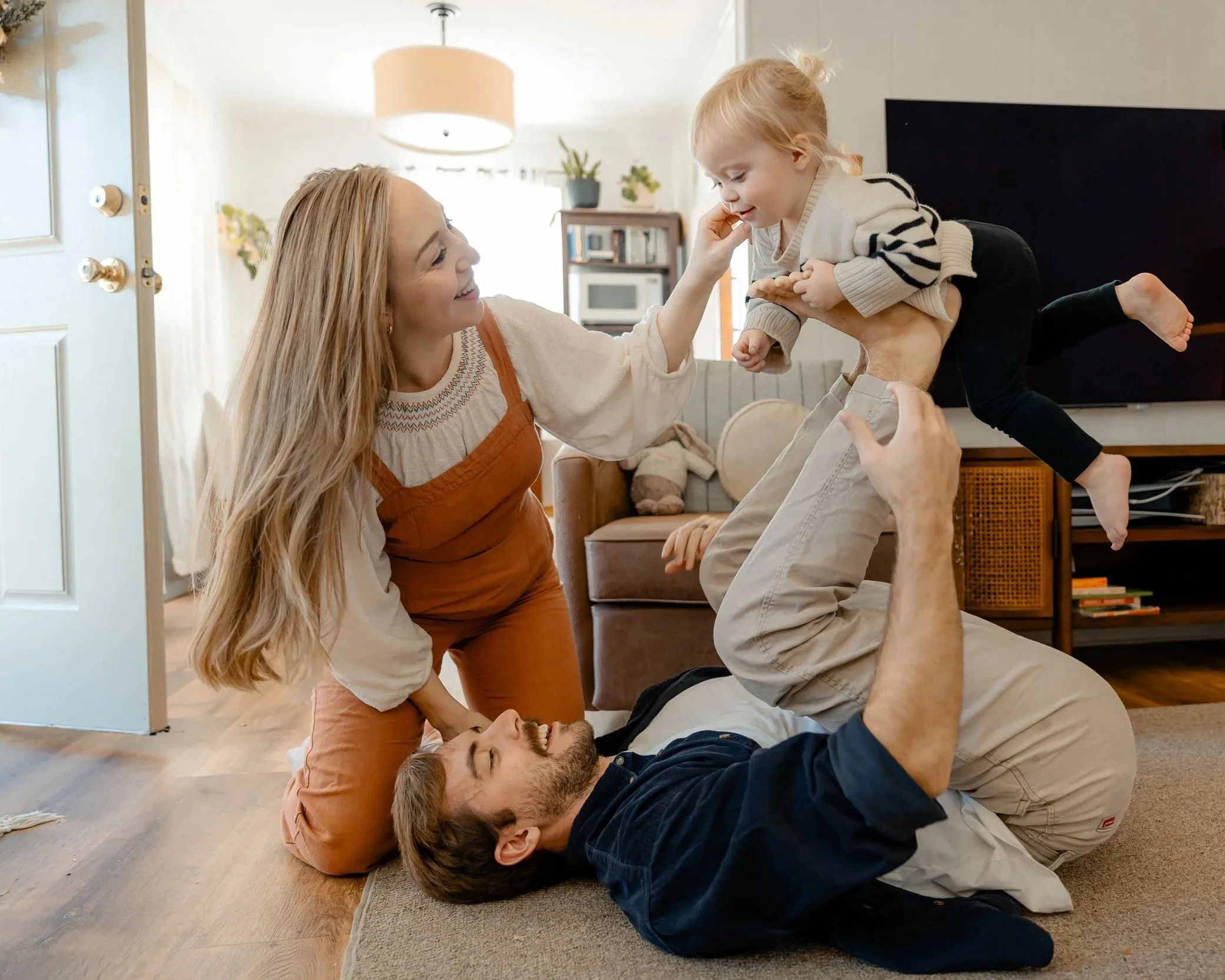 A smiling family of four playing on the living room floor, with a woman, man, young girl, and boy lifting and playing.