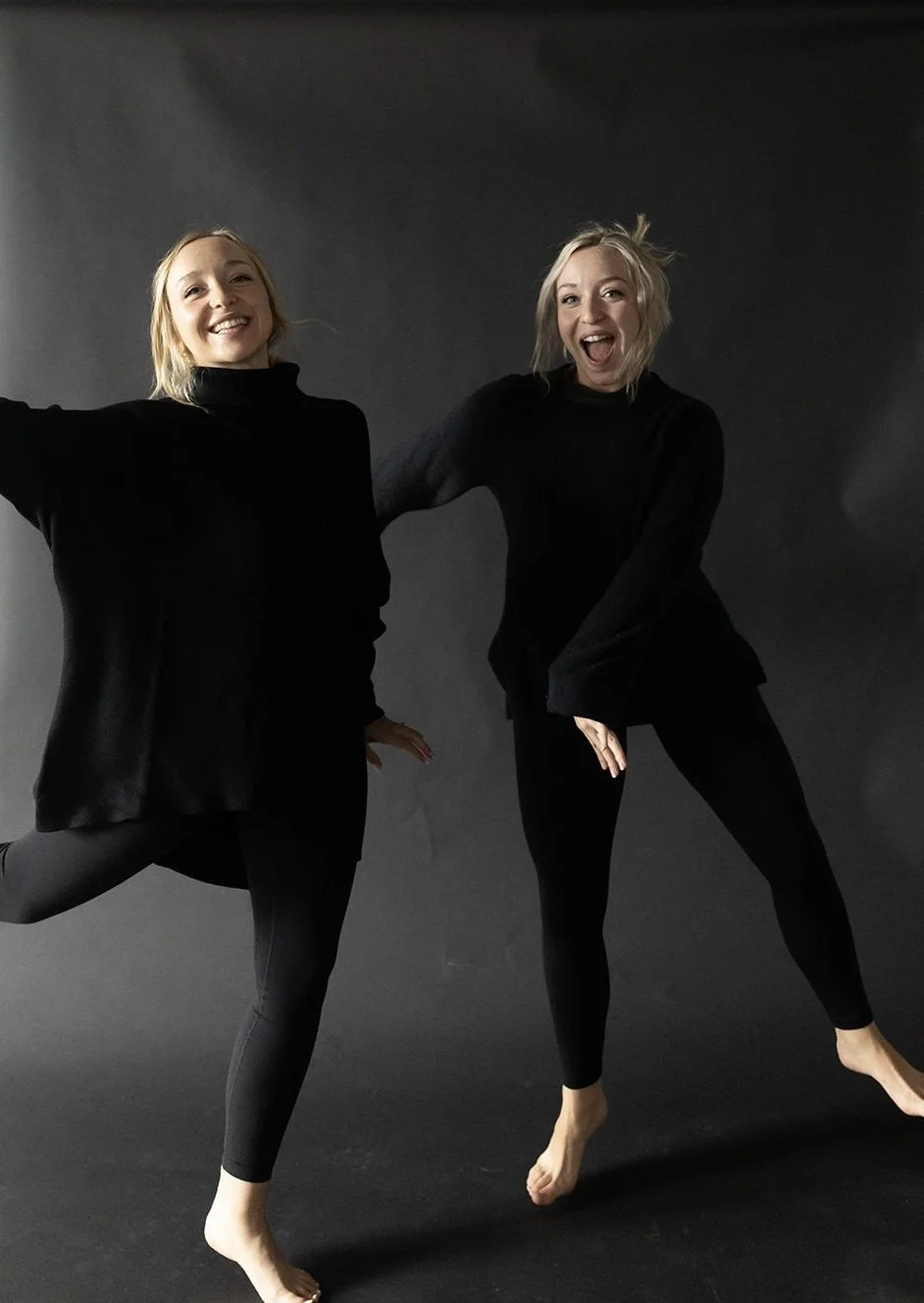Two women in black outfits posing happily against a dark background, one with arms outstretched and both with their feet off the ground.