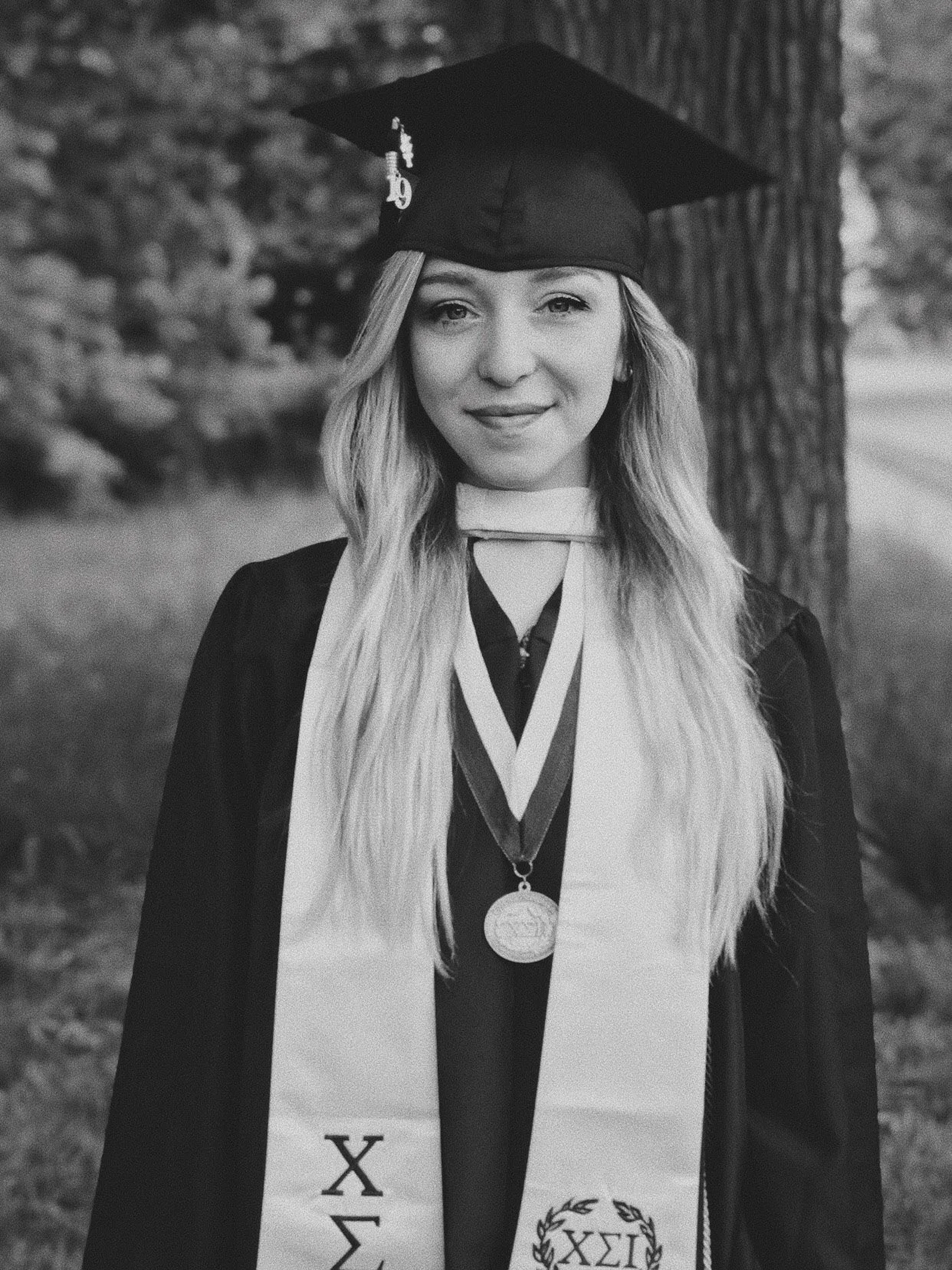 A young woman in a graduation cap and gown, wearing a medallion and a sash, standing outdoors near a tree.