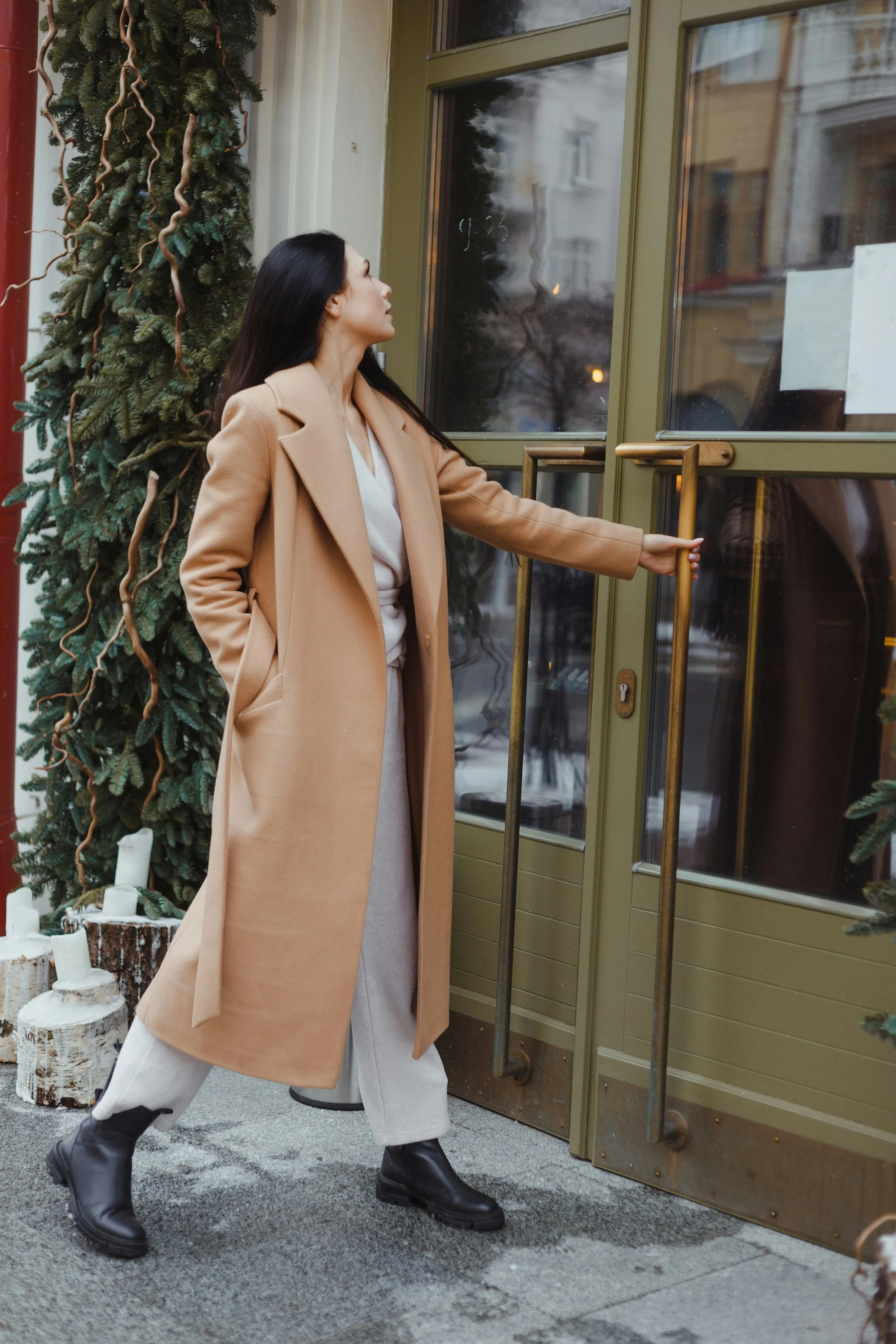 Woman in a camel coat and gray pants opening a green door with gold handles outside a building decorated with Christmas greenery