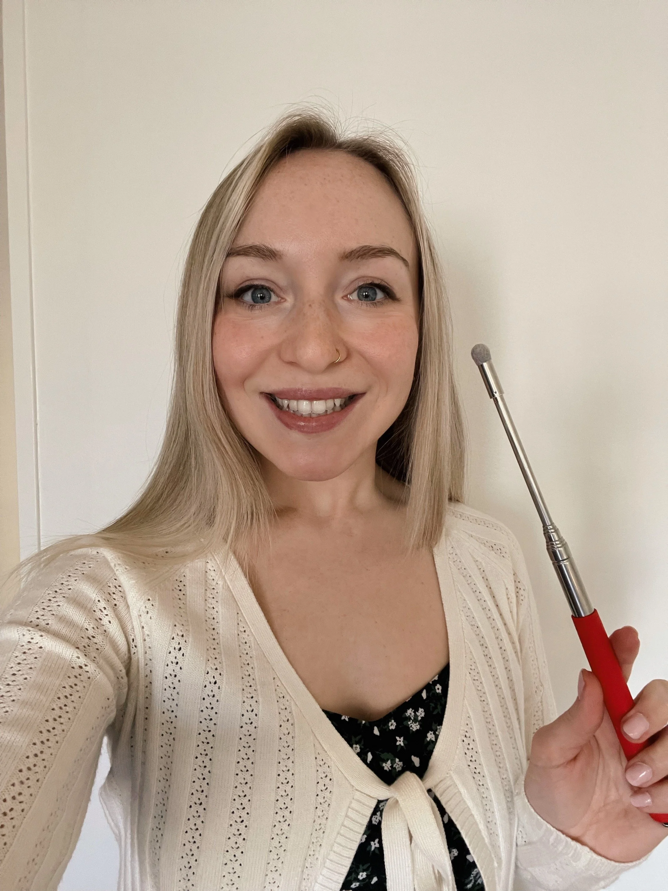A woman with blonde hair, blue eyes, and a nose ring smiling and holding a small brush.
