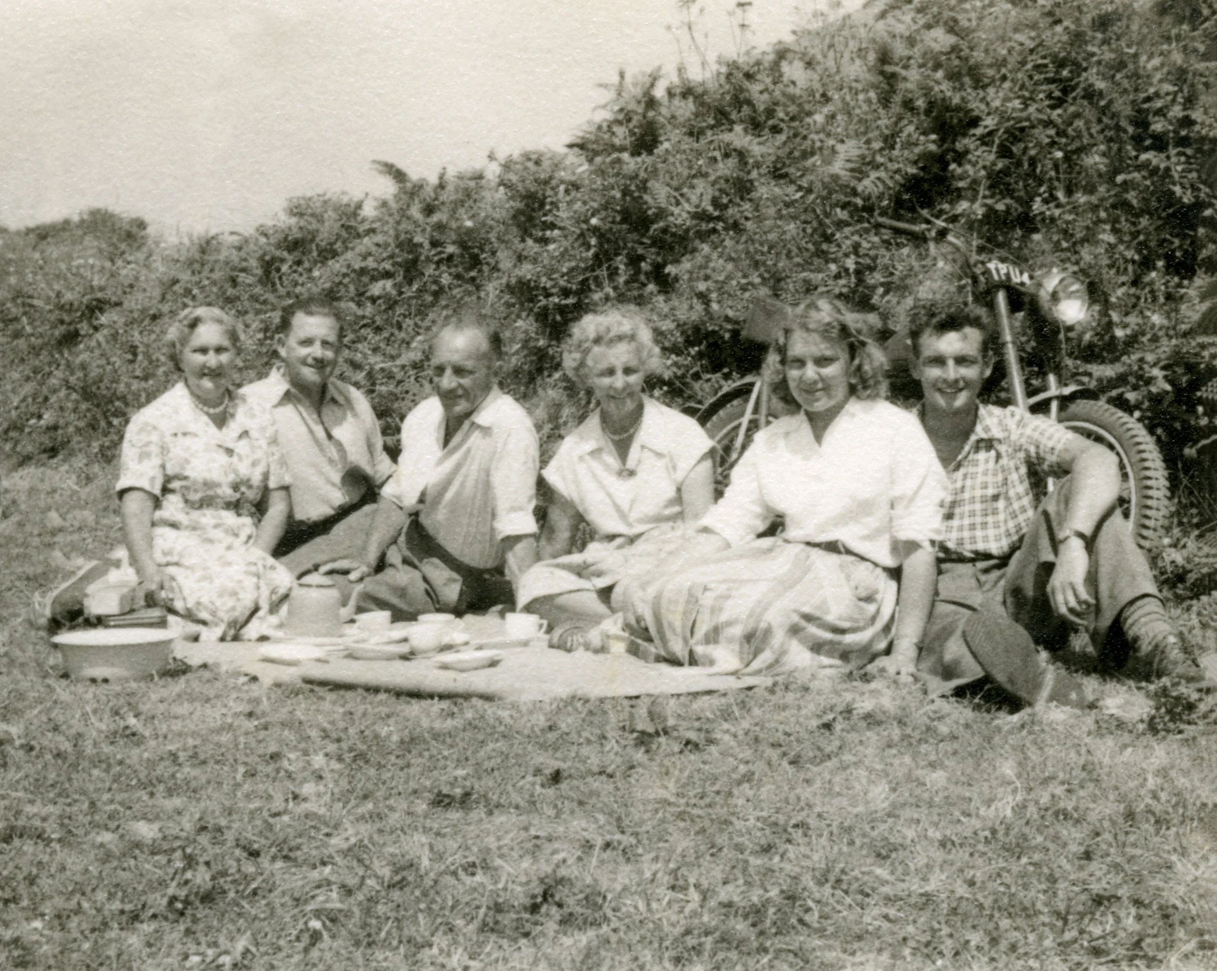 Black and white photo of seven people sitting on grass outdoors, enjoying a picnic, with a vintage motorcycle in the background.