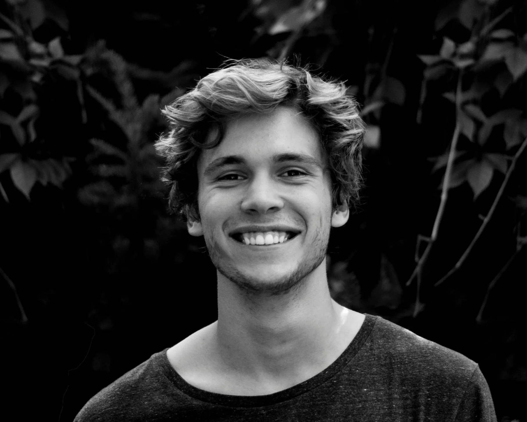 Black and white portrait of a young man with curly hair, smiling, wearing a dark t-shirt, with foliage in the background.
