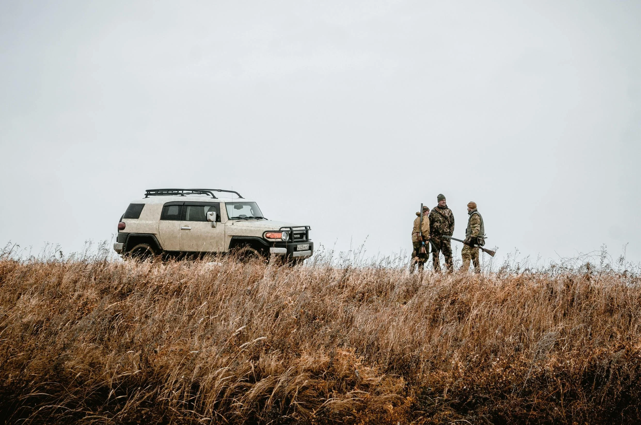 Three hunters in camouflage and outdoor clothing stand in a grassy field with a beige SUV parked nearby under an overcast sky.