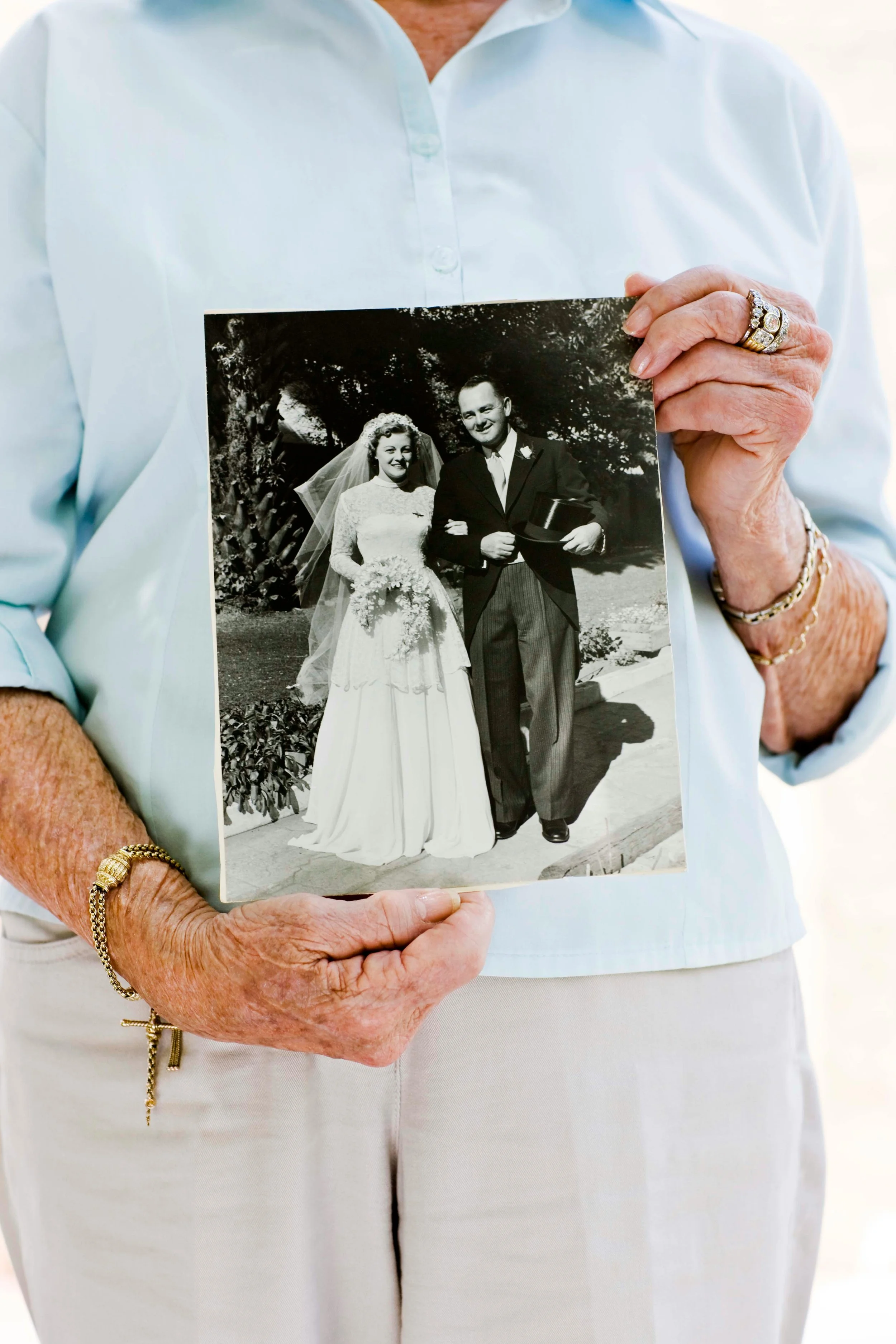 An elderly woman wearing a light blue blouse holds a black-and-white wedding photo of a bride and groom outdoors.