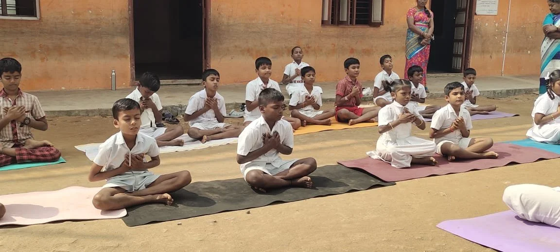 Children sitting cross-legged on mats outdoors, meditating or praying, with two women standing behind them.