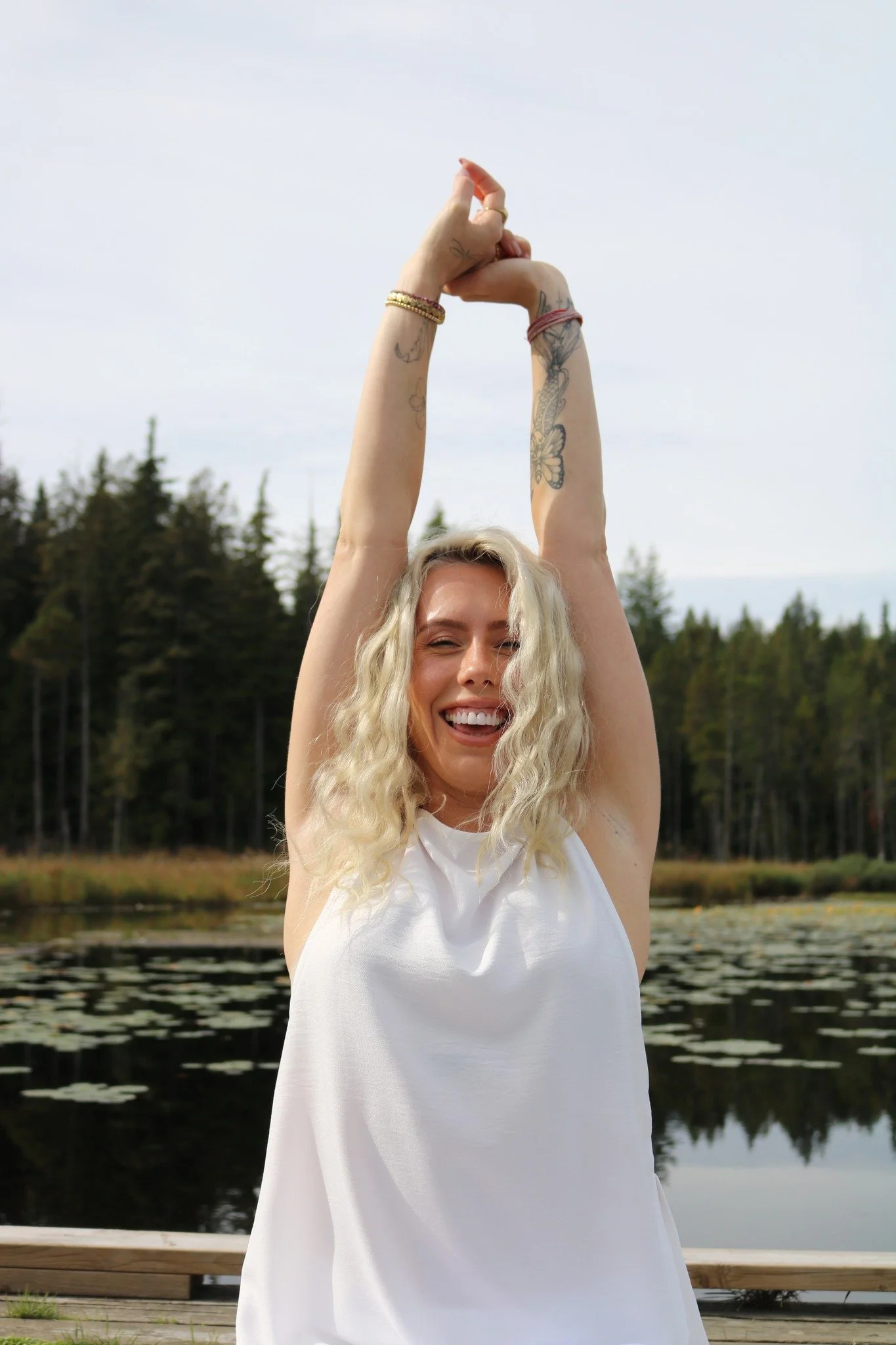 A woman with blonde curly hair smiling and raising her arms above her head, forming a gesture with her fingers, outdoors with trees and water in the background.