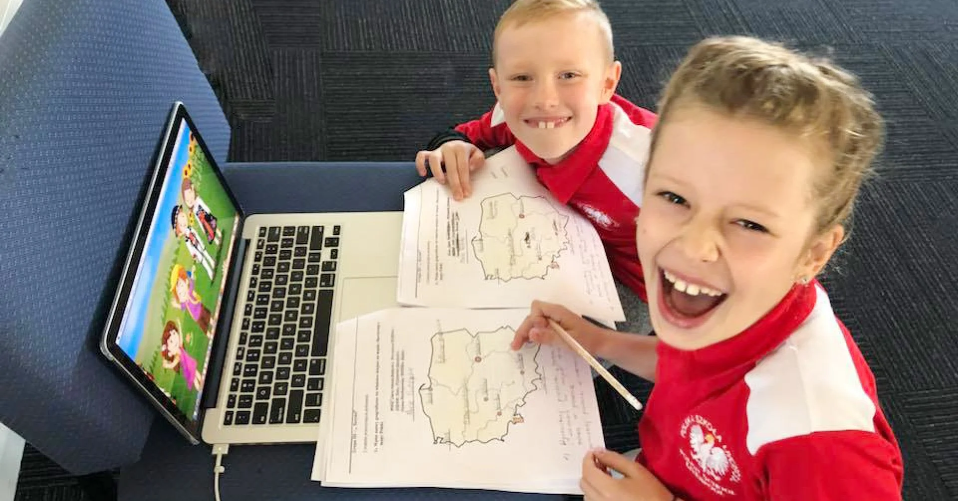 Two young boys in red and white shirts are sitting at a table with a laptop and open workbooks. They are smiling and looking up at the camera. The laptop screen shows animated characters in a colorful outdoor scene.