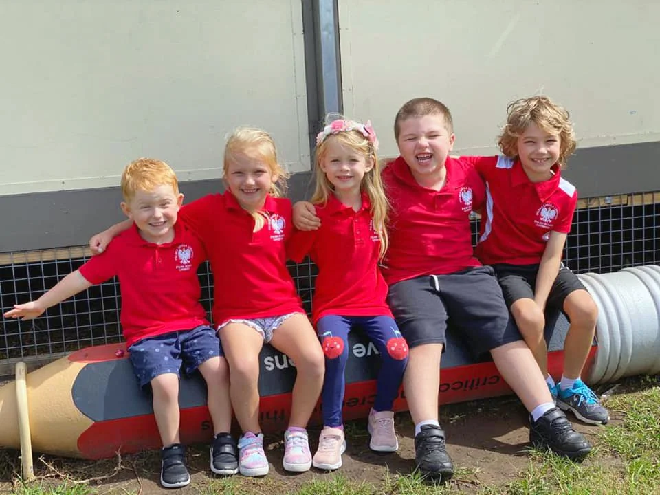Group of five young children sitting together on a large outdoor model rocket, all wearing matching red shirts, smiling and posing for a photo.