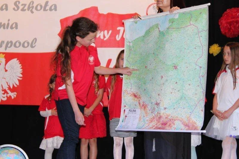 Group of children with a teacher, pointing at a large map of Poland, during a presentation or school event.