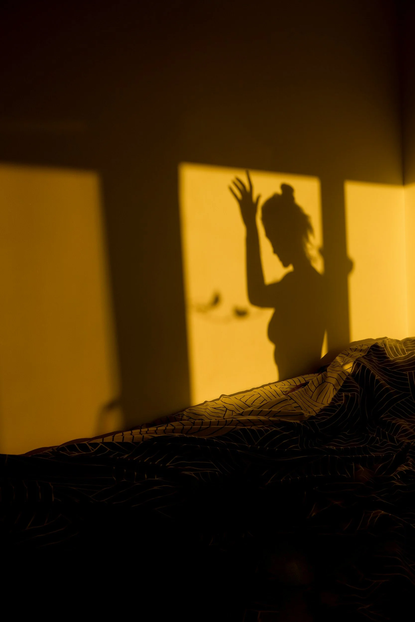 Shadow of a person with their hand raised, cast on a wall illuminated by warm yellow light, with a bed and striped blanket in the foreground.