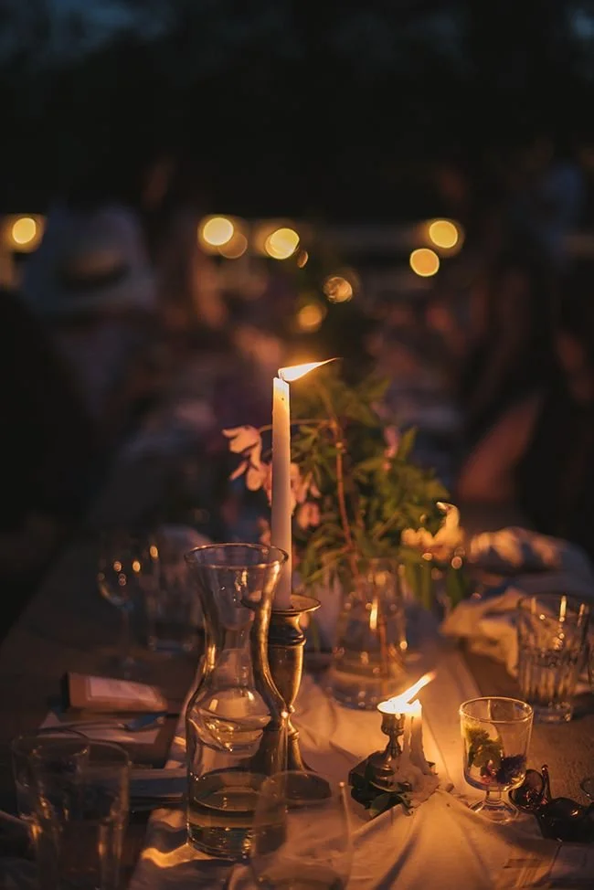 A table set outdoors at night with candles, glasses, and flowers, illuminated by candlelight with blurred background of people and string lights.