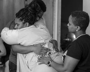 Two women embrace in a hug while a woman on the right looks on, in a room with a door and an outside scene visible in the background, in black and white.