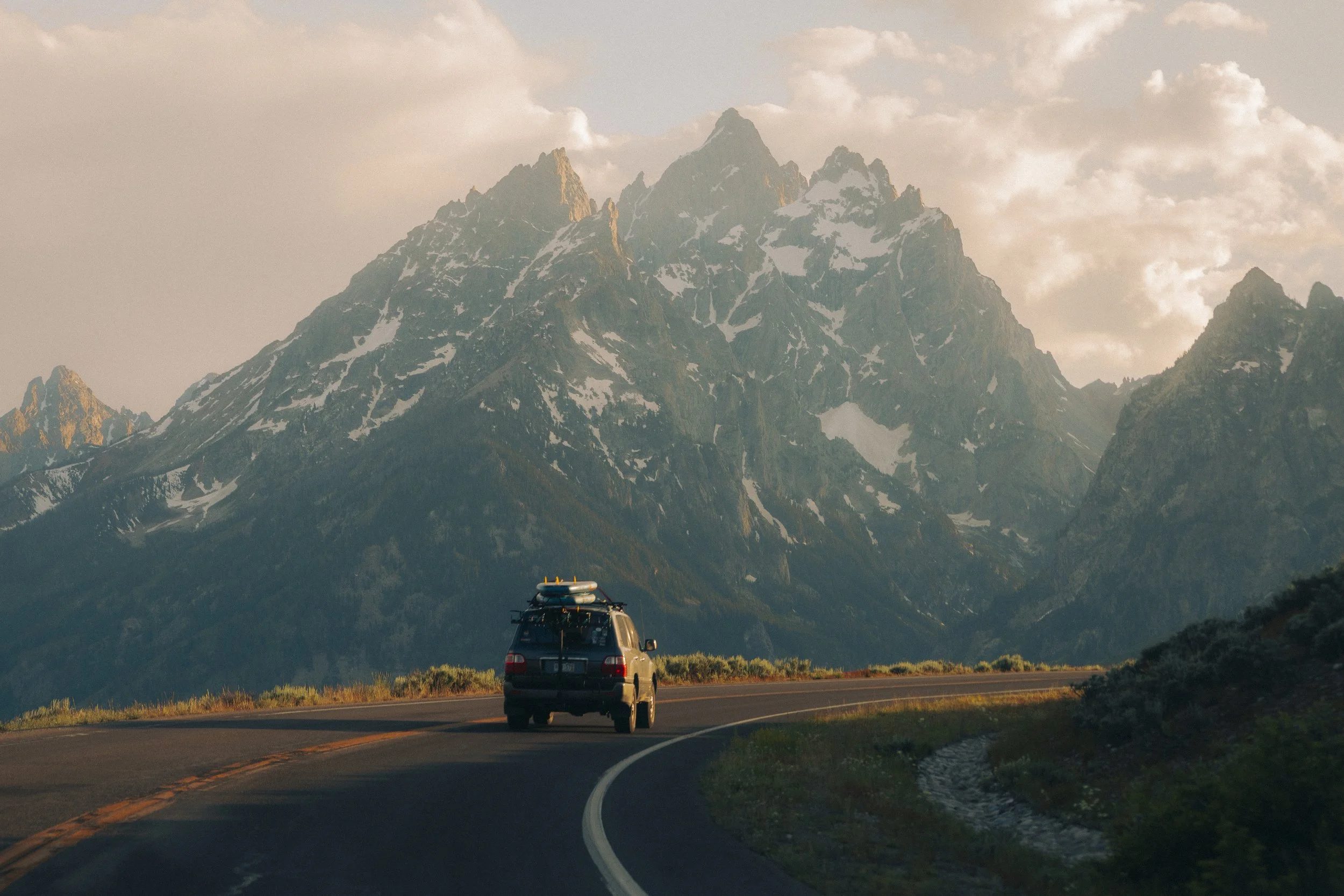 An SUV driving on a curvy mountain road with snow-capped peaks in the background during sunset.