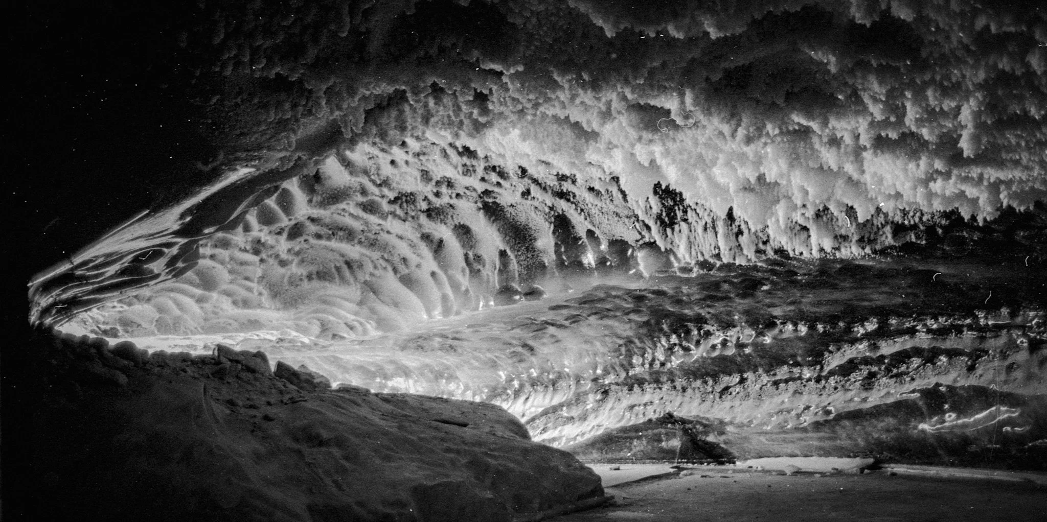 A black and white photo of an ice cave with a large, textured, icy ceiling and formations on the walls, and snow and ice on the ground.