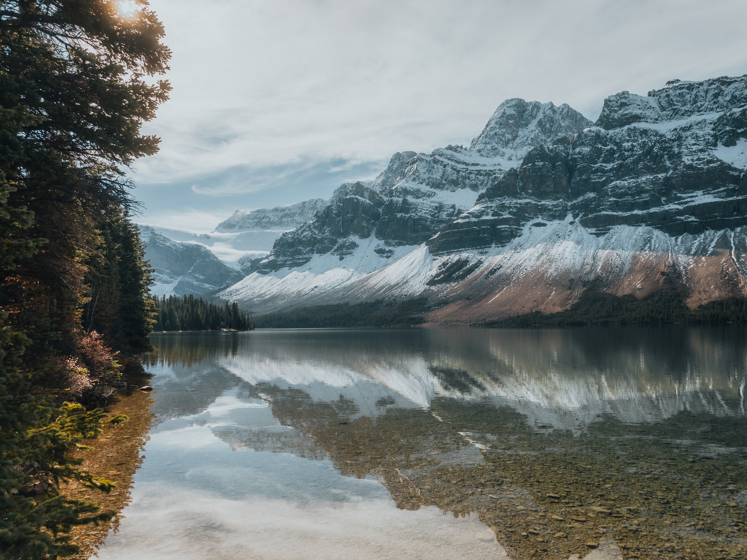 Snow-capped mountains reflected in a calm lake with trees along the shoreline under a cloudy sky.
