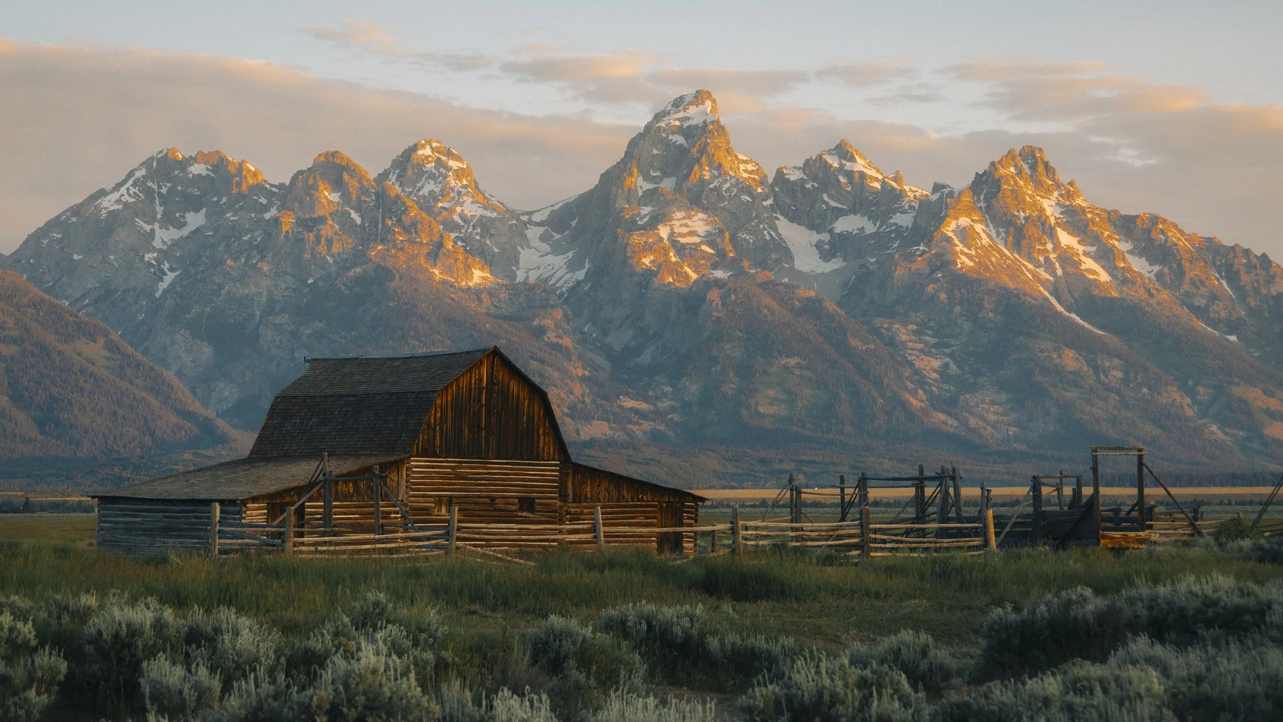 A rustic wooden barn in a green field with mountain peaks in the background, bathed in warm sunlight.