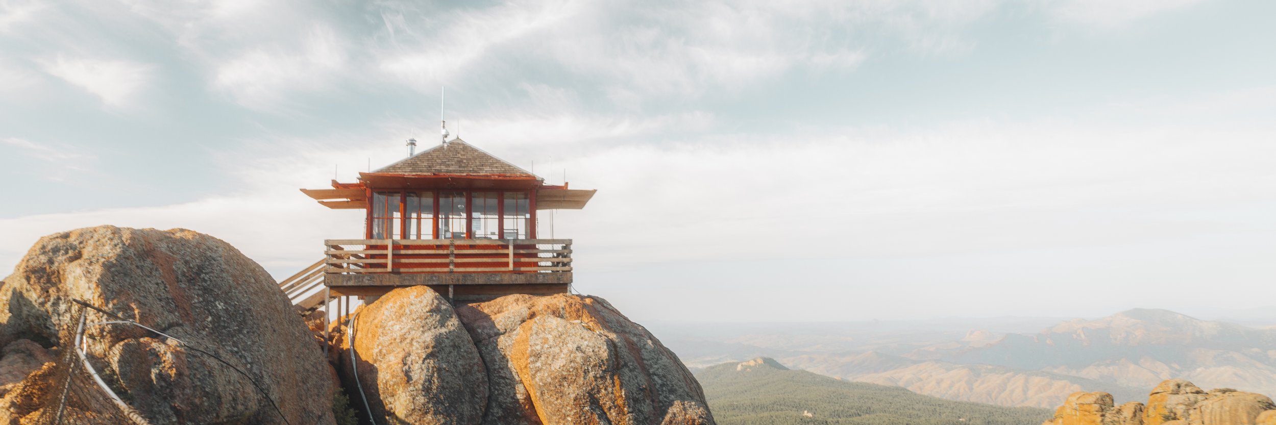 A wooden lookout tower on a rocky mountain peak with a mountainous landscape in the background, under a partly cloudy sky.