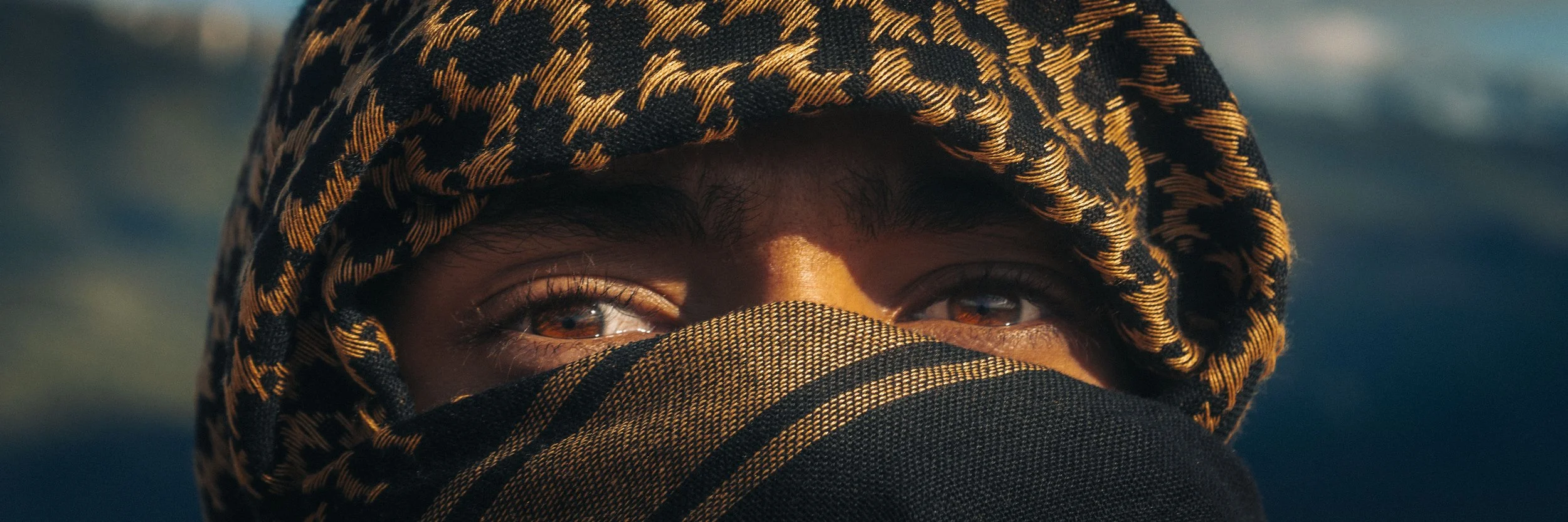 Close-up of a person's face with brown eyes, wearing a black and orange patterned headscarf and a black mask covering the bottom part of the face.