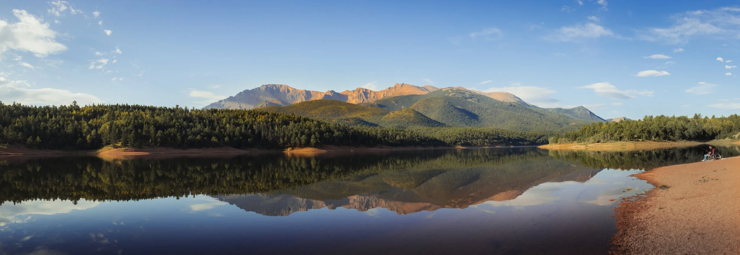 A calm body of water reflecting the surrounding trees and distant mountains under a blue sky with some clouds.
