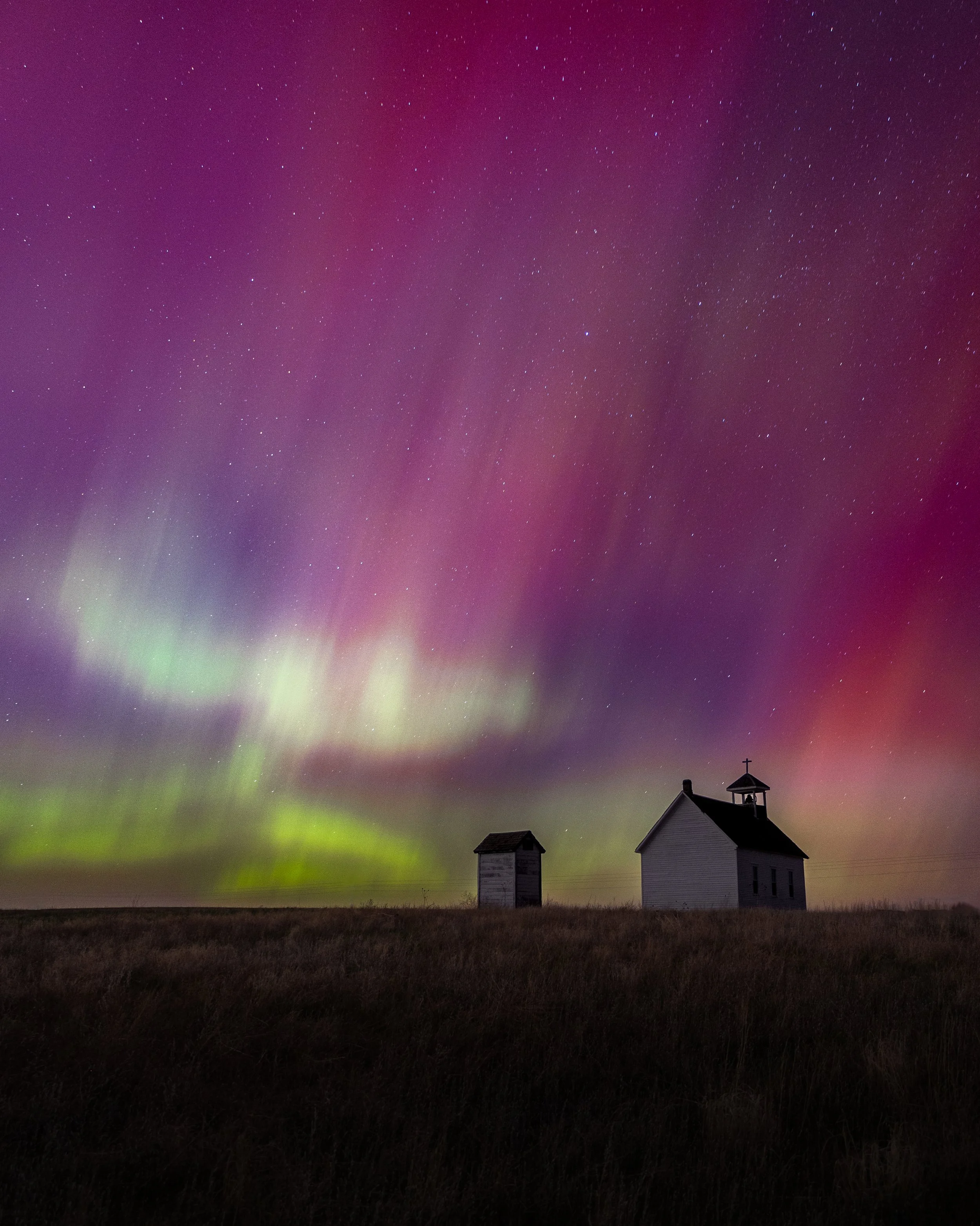 Night sky with colorful aurora borealis over two small buildings, a barn and a church, in a field.