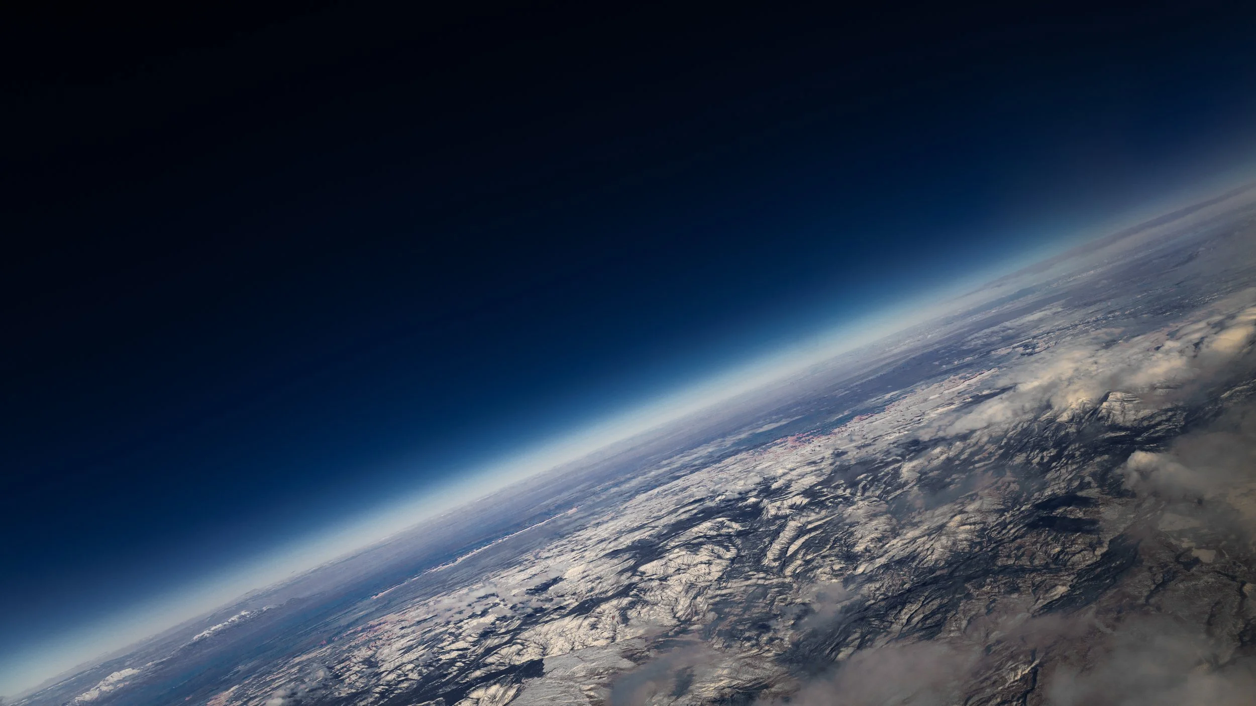 View of Earth from space showing a curved horizon, with snow-covered mountains and clouds over land.