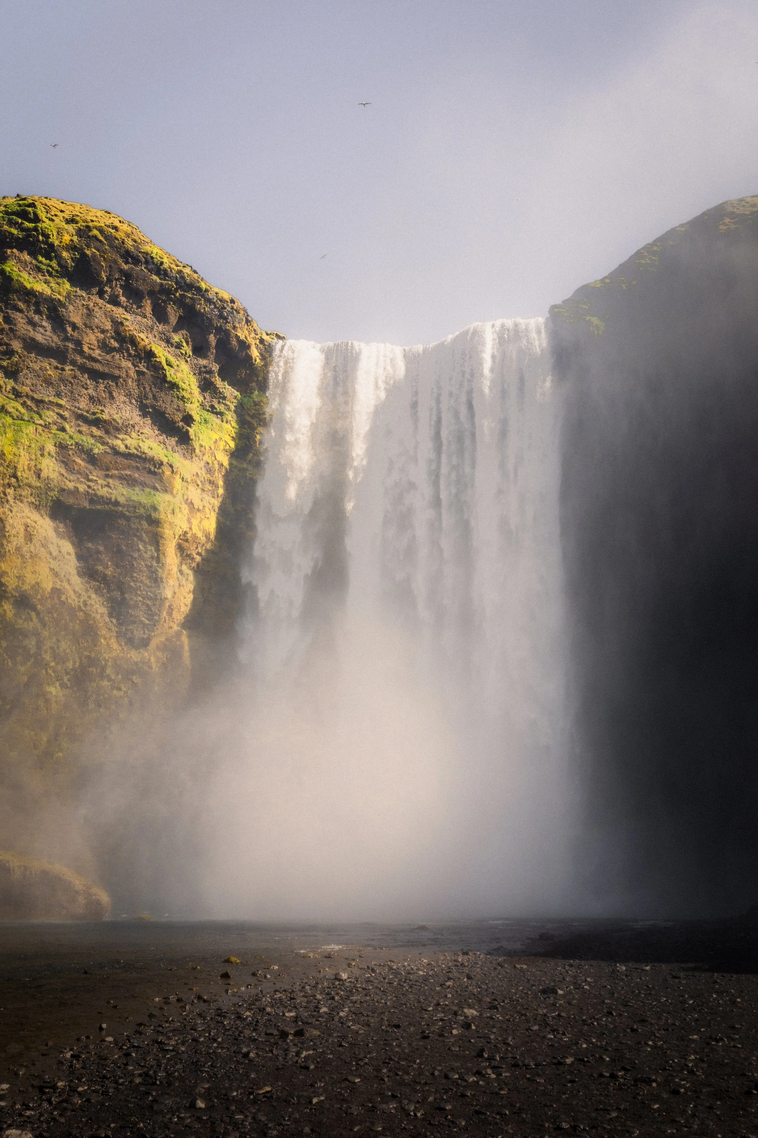 A large waterfall cascading down moss-covered cliff sides with mist rising from the base, and a few birds flying in the sky.