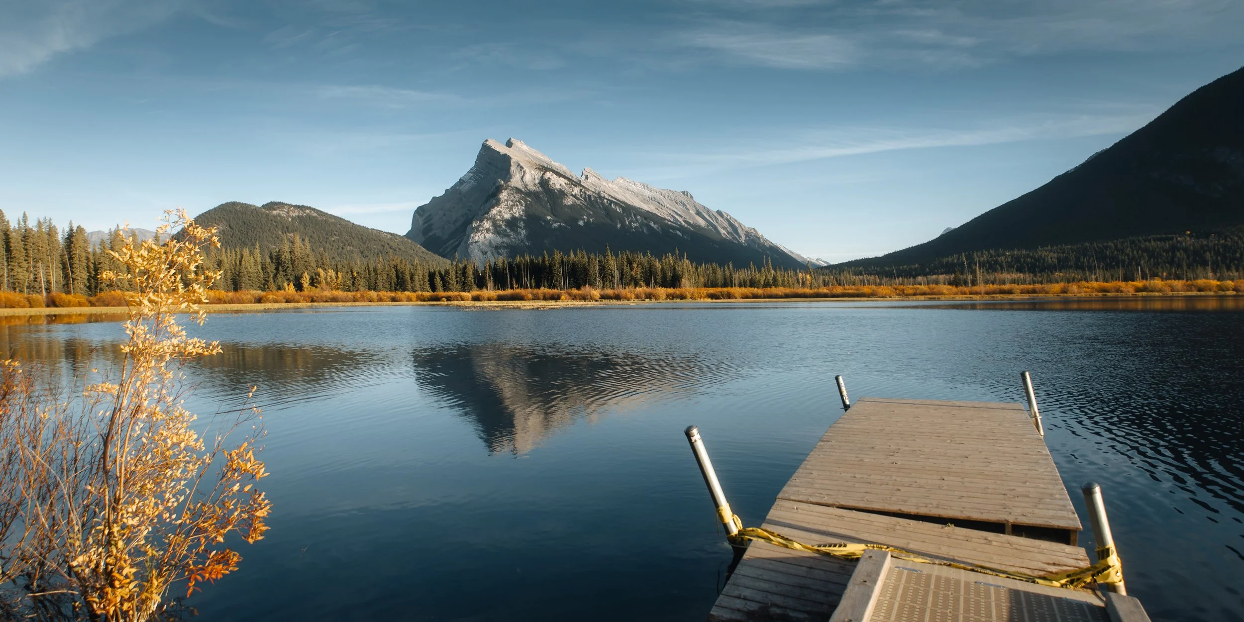 A mountain lake with calm water reflecting the surrounding forest and a snow-capped mountain in the background. There is a wooden dock extending into the lake in the foreground.