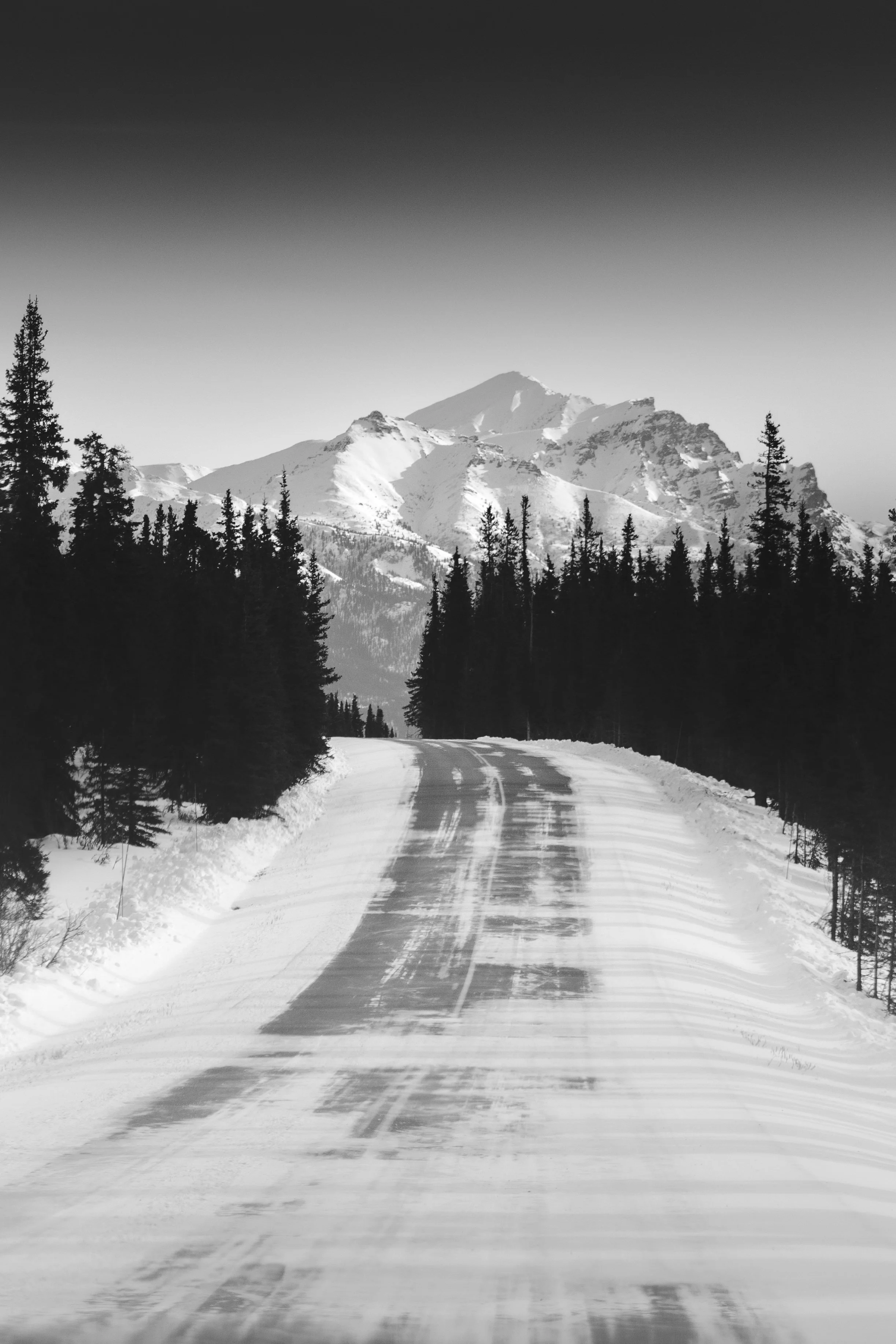 Snow-covered mountain road passing through a forest with snow-covered peaks in the background, black and white photograph.