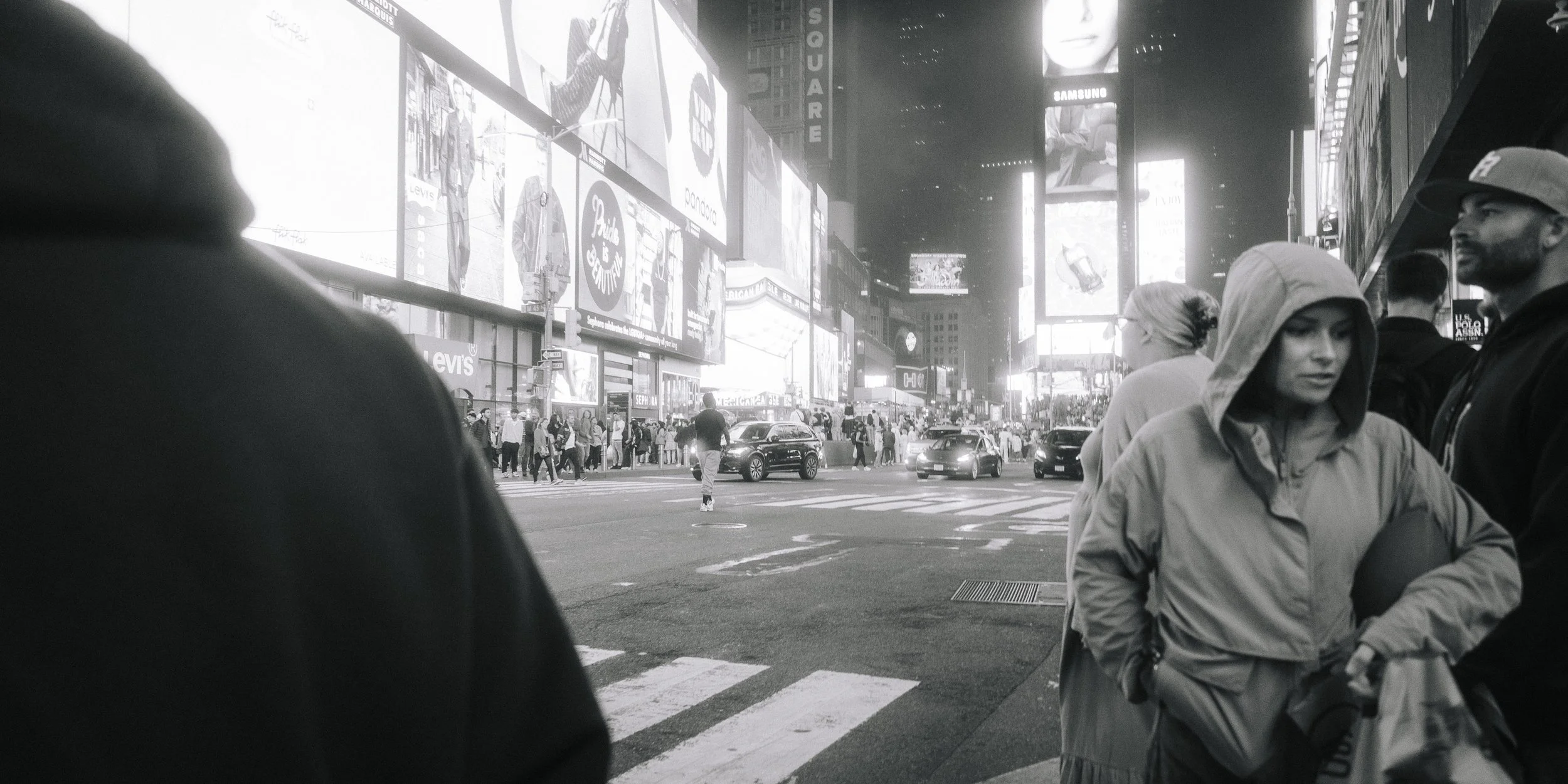 Nighttime city street scene with bright digital billboards, people waiting to cross the crosswalk, and vehicles on the road.