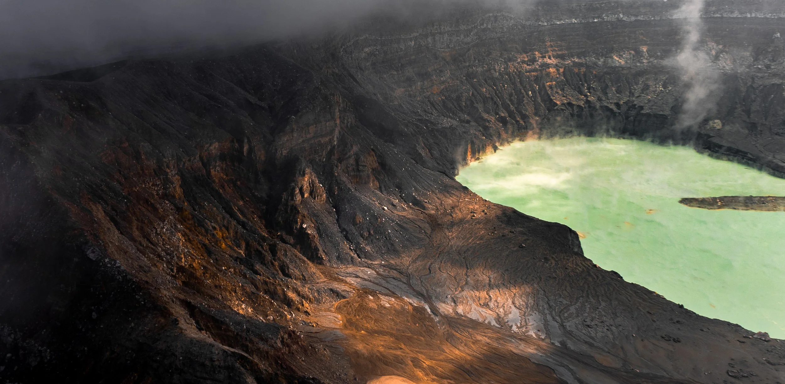 A volcanic crater with dark, rugged slopes and a greenish lake at the bottom, with steam rising from the lake.