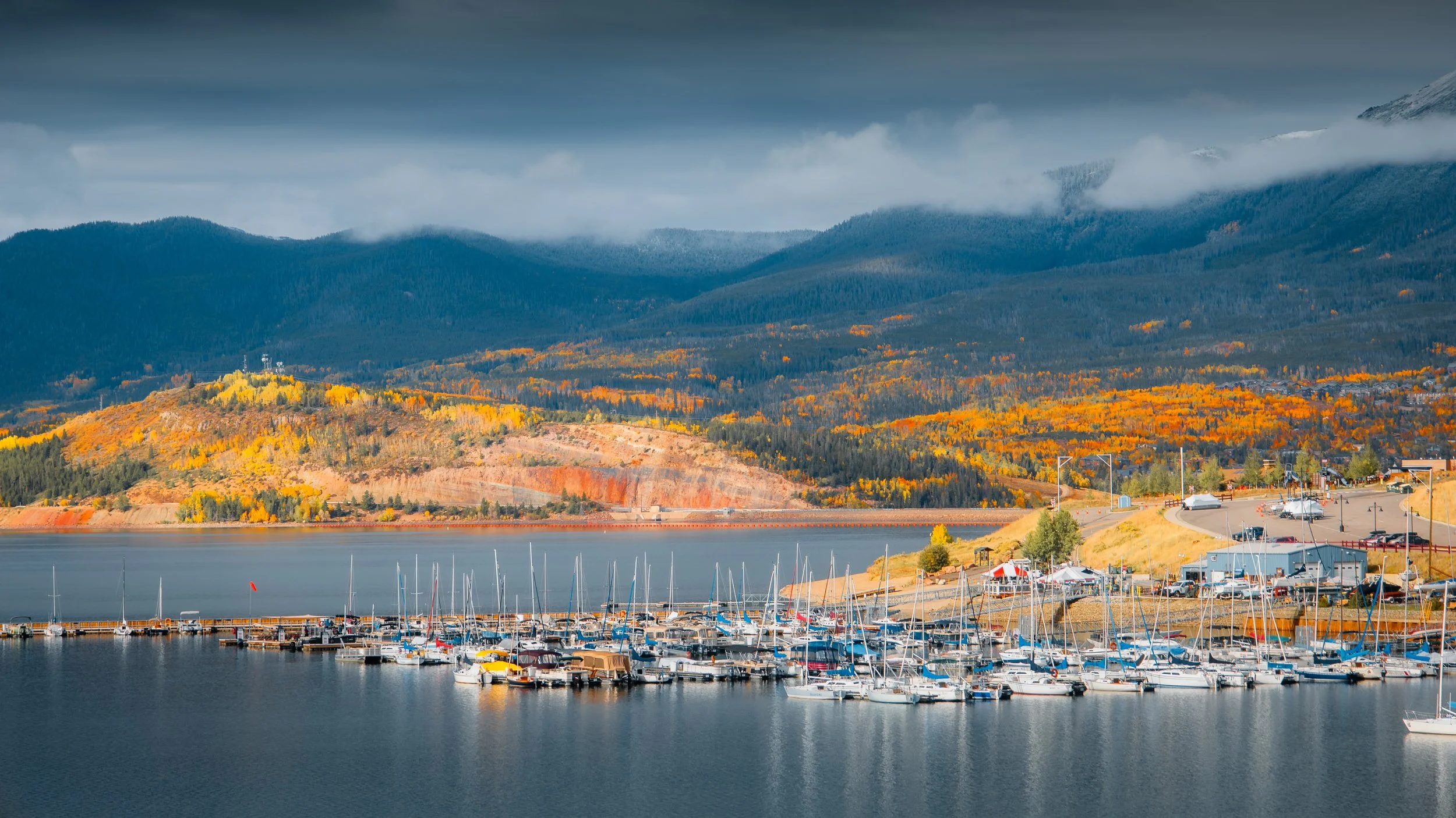 A scenic view of a marina with numerous sailboats and yachts docked, set against a backdrop of colorful autumn trees, rolling hills, and mountains under a cloudy sky.
