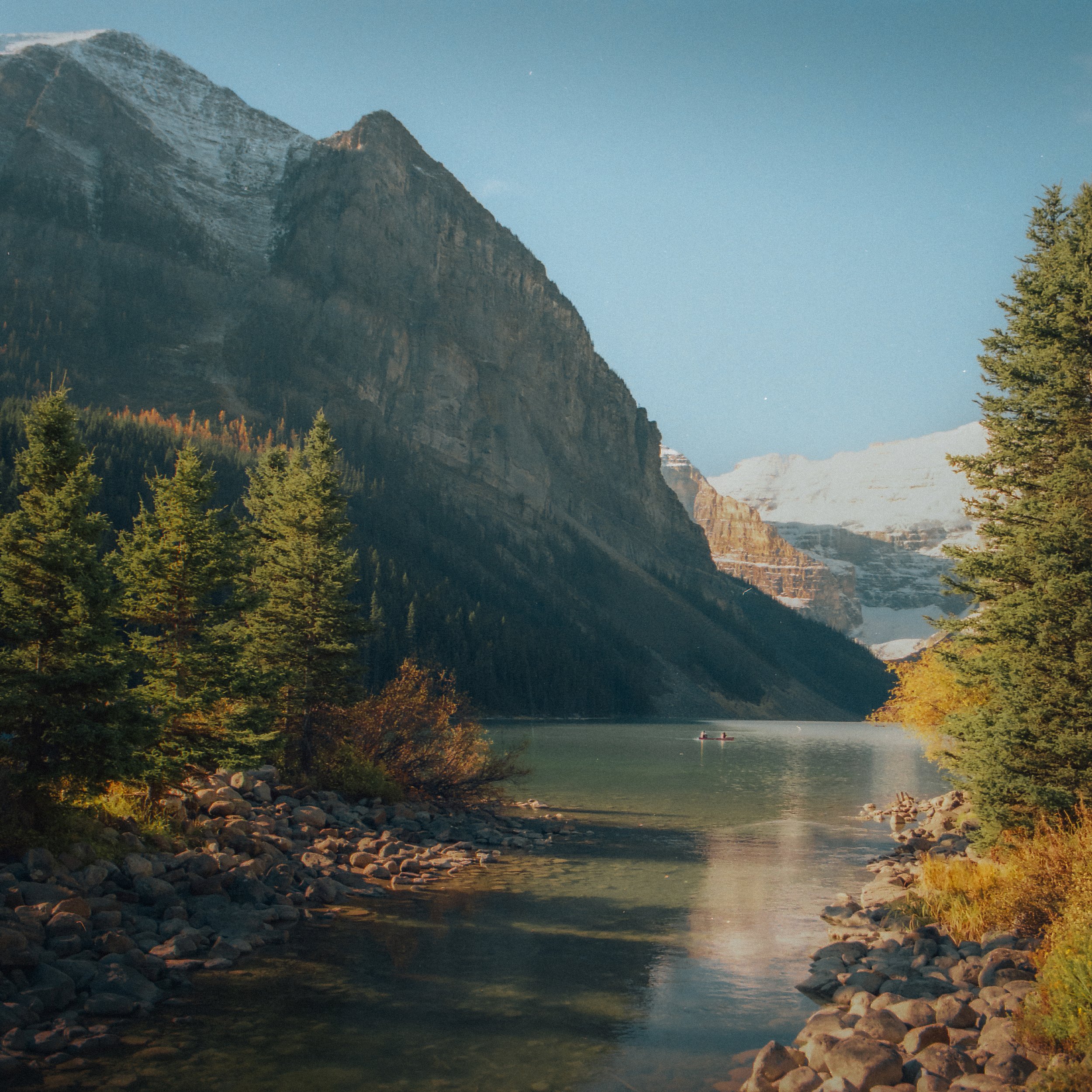 A scenic landscape of a lake surrounded by tall mountains and dense evergreen trees, with a clear blue sky overhead.