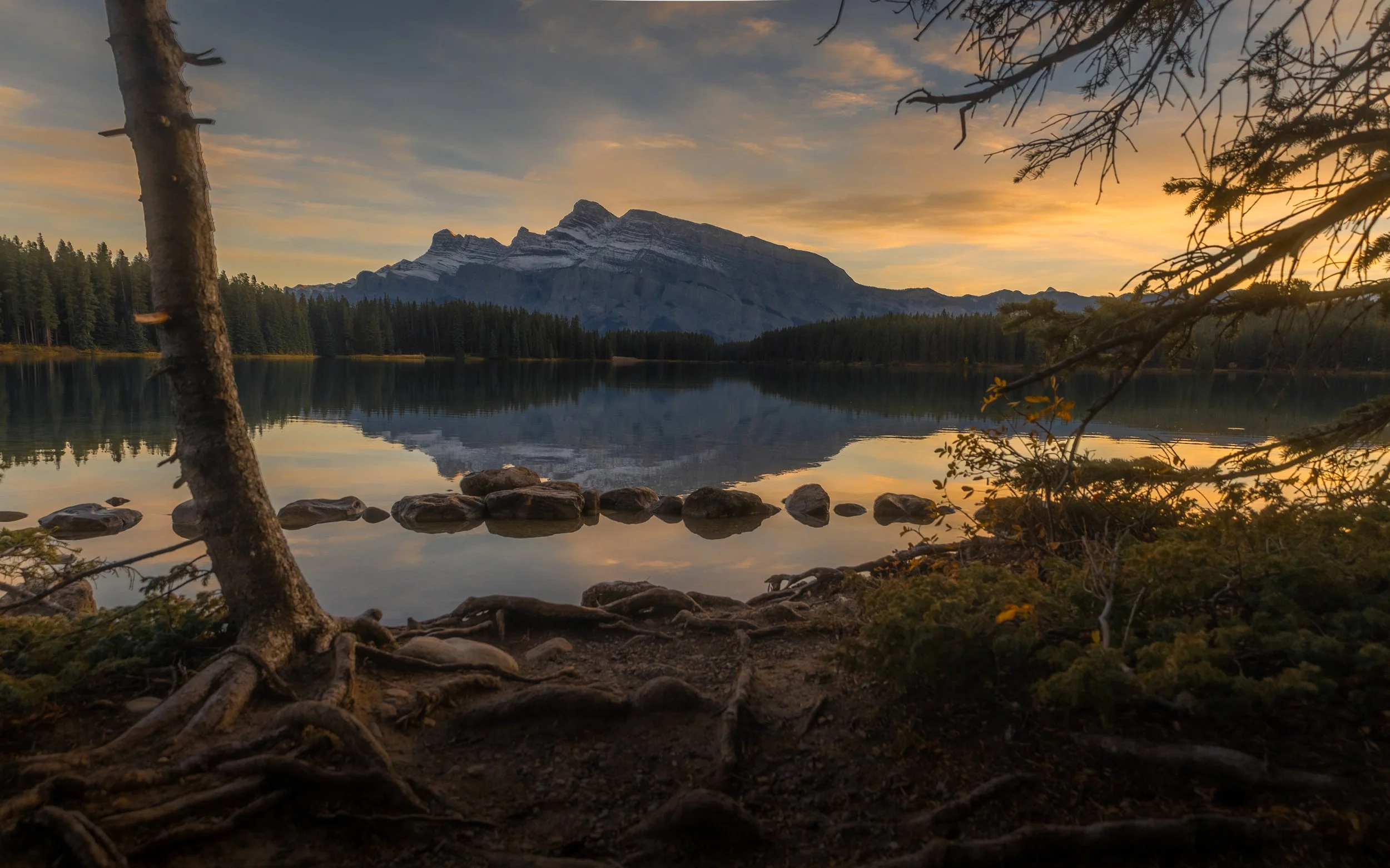 A peaceful mountain lake scene at sunset with a mountain range in the background, a forest at the lake's edge, and rocks in the water, framed by trees and branches.