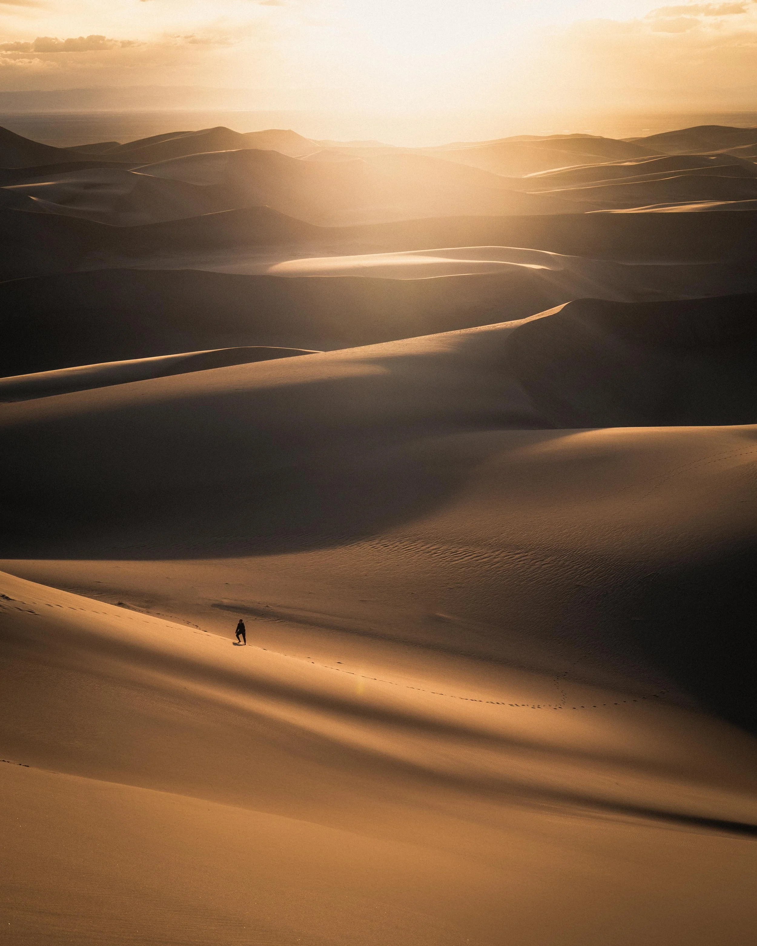 A person walking alone across sand dunes in a desert at sunset with warm, golden lighting and rolling dunes extending to the horizon.