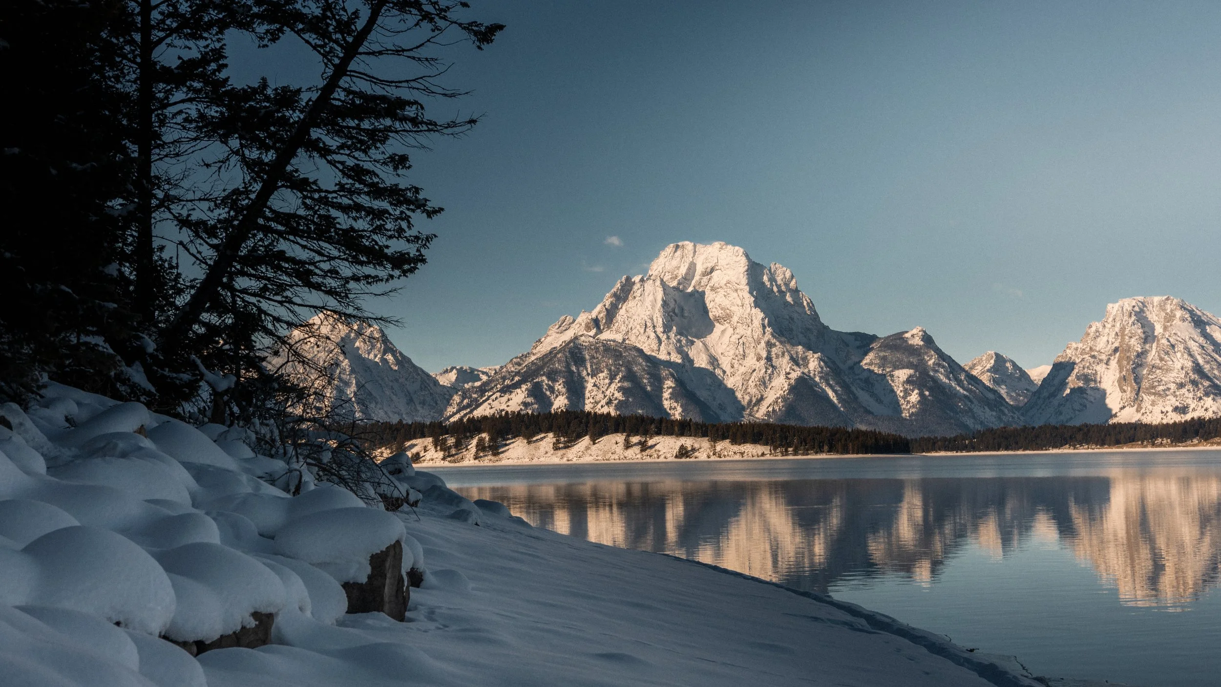 Snow-covered shoreline with trees in the foreground and a mountain range reflected in a calm lake under a clear blue sky.