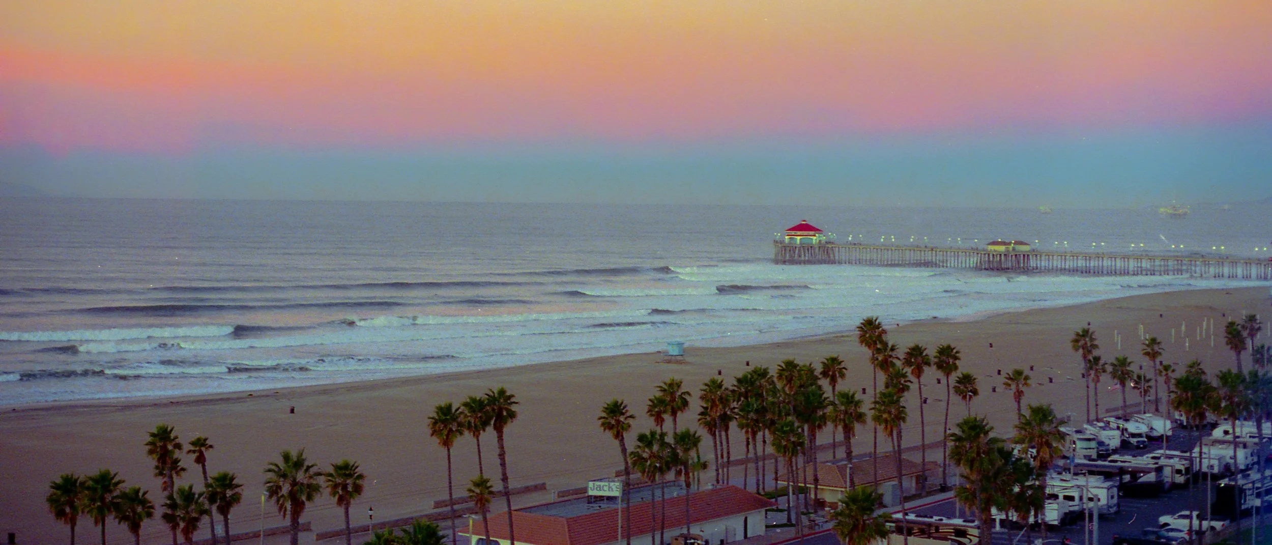Sunset over a sandy beach with palm trees, a pier extending into the ocean, and boats on the water in the background.