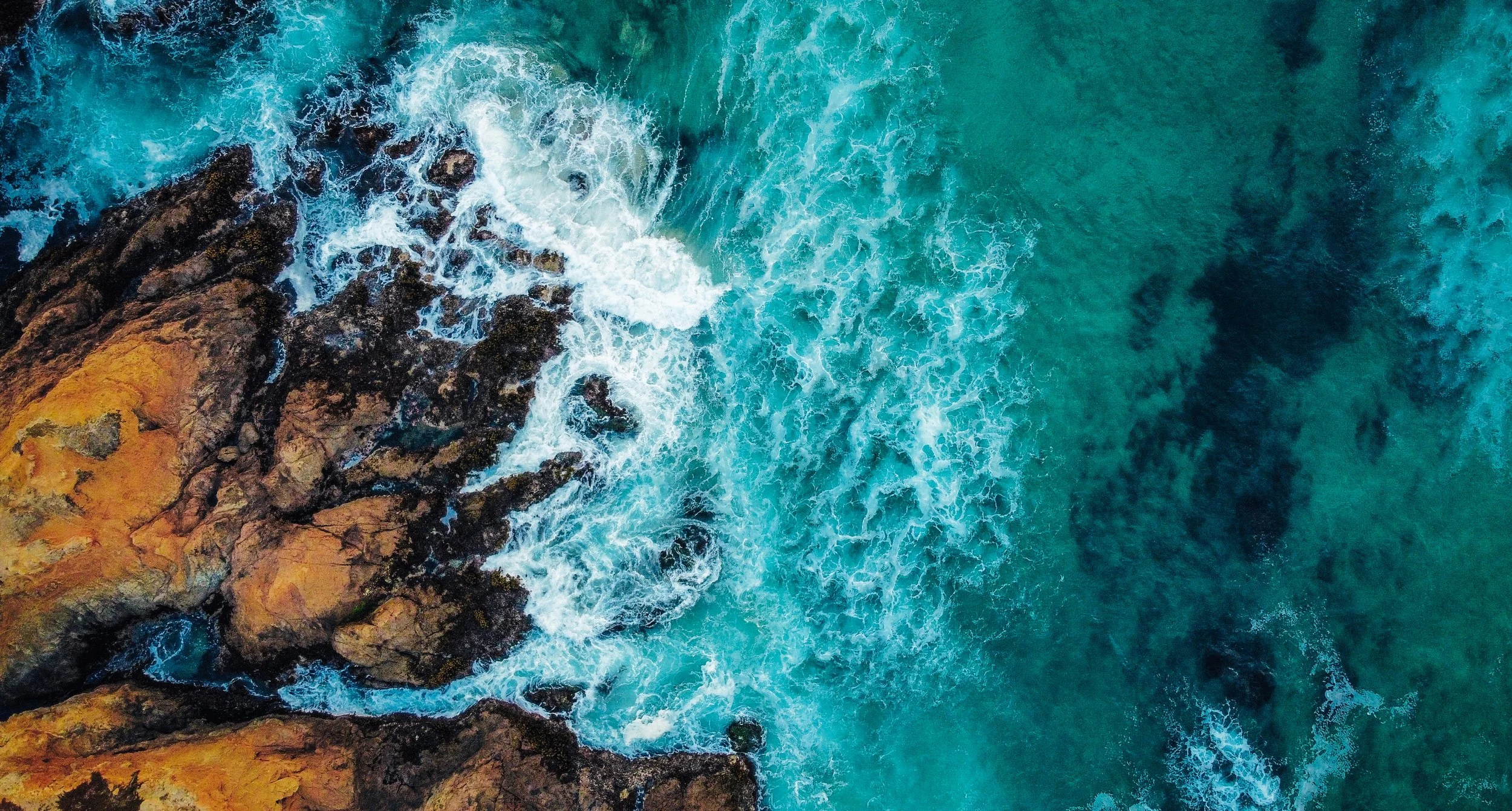 Aerial view of teal ocean waters crashing against rocky shoreline with yellow and dark brown rocks.