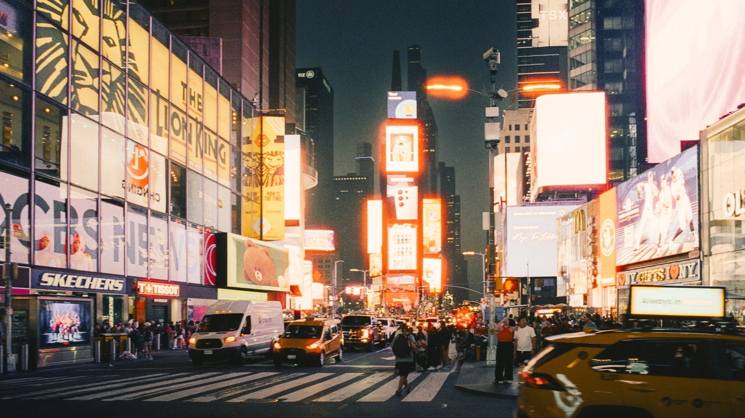 Nighttime view of Times Square in New York City with bright digital billboards, cars, and people crossing the street.