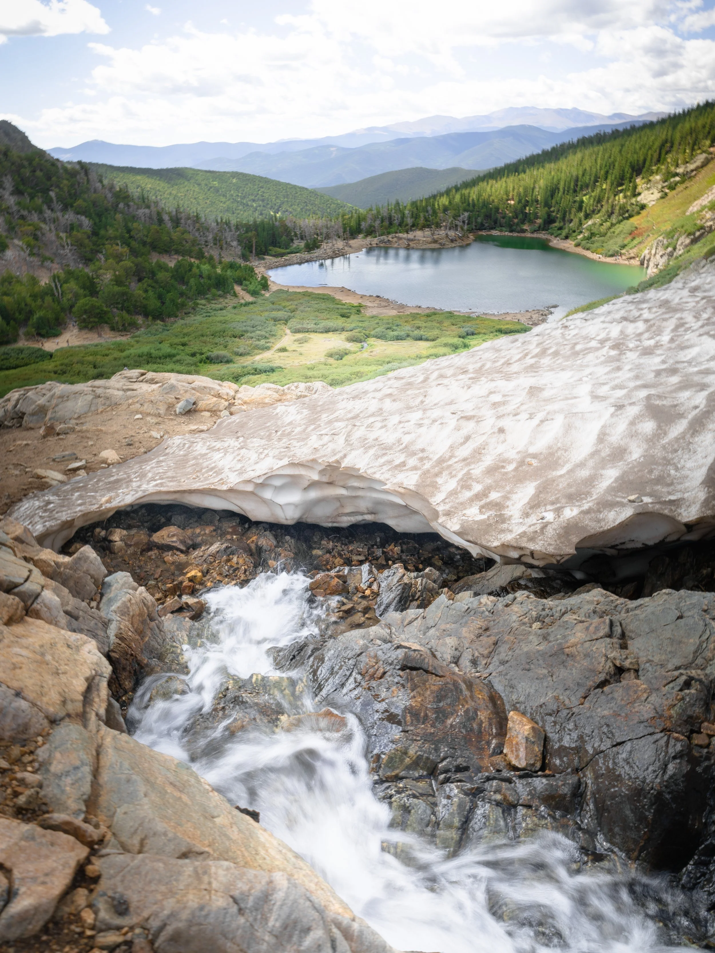 Mountain stream flowing over rocks with snow in the foreground, and a lake surrounded by forested mountains in the background under a cloudy sky.