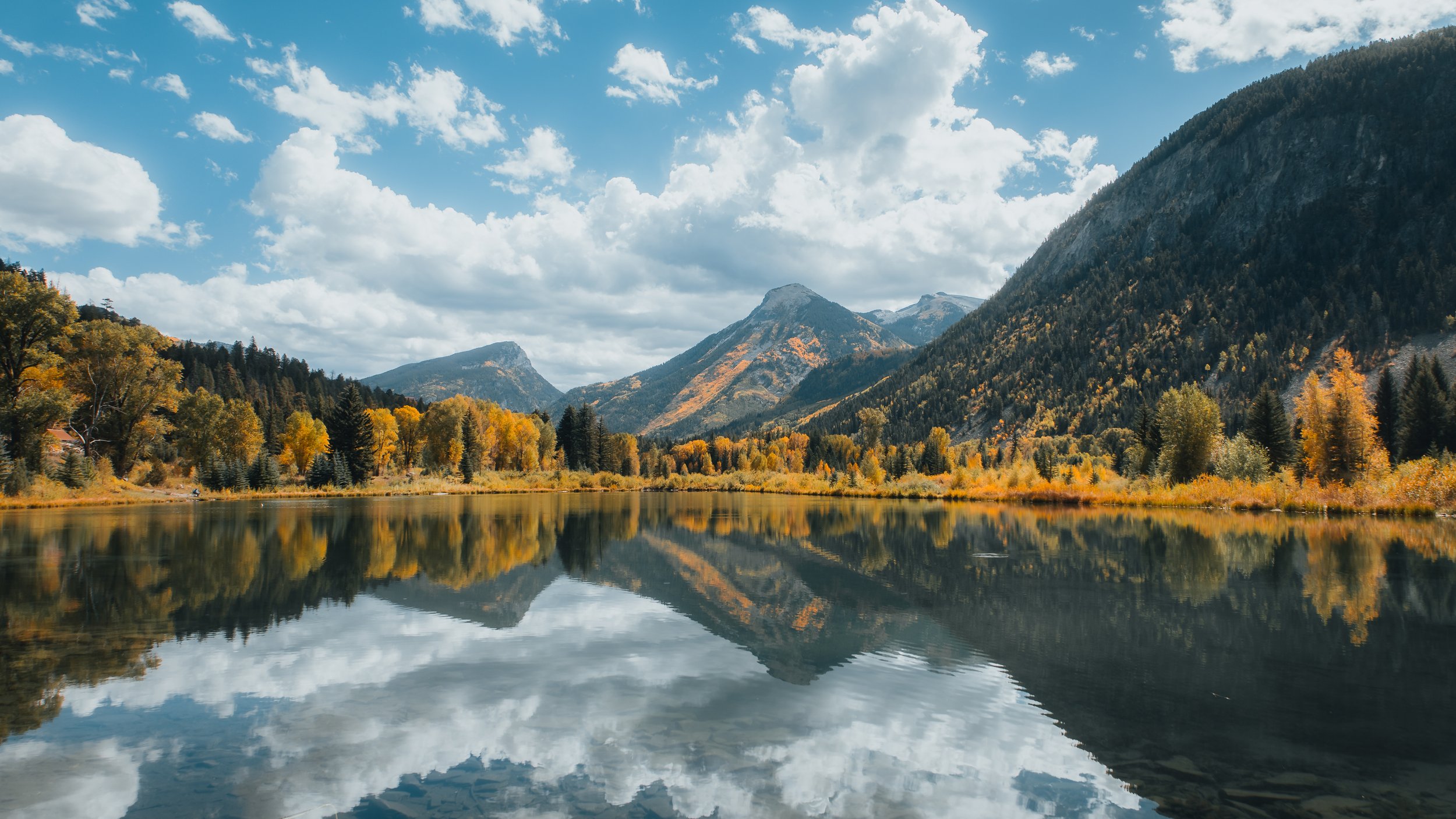 Scenic landscape of mountains, trees with fall foliage, and a calm lake reflecting the sky and mountains under a partly cloudy sky.