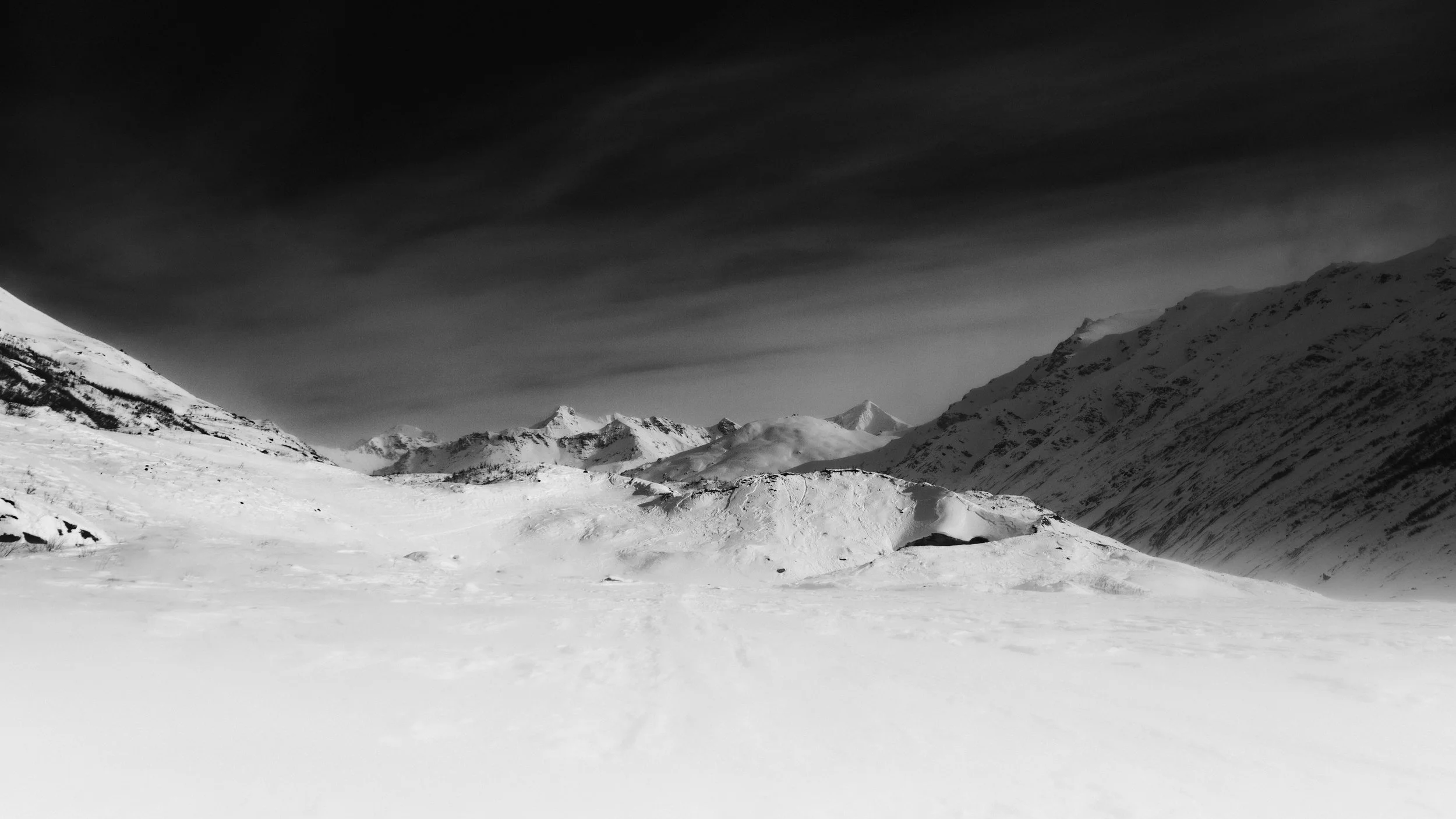 Snow-covered mountain landscape with dark sky, distant peaks, and ridges.