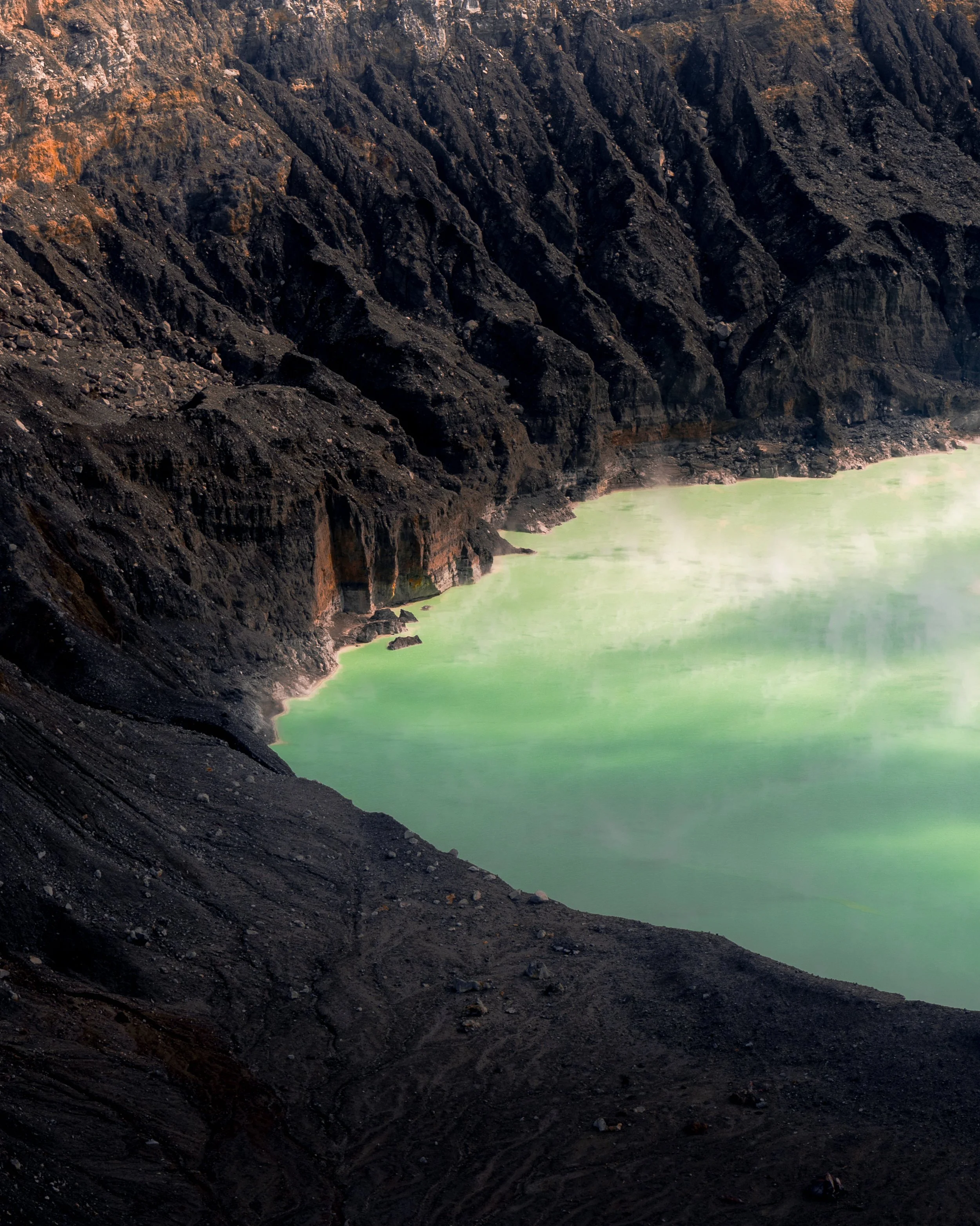 A volcanic crater with greenish water and dark rugged rocky edges, with steam rising from the surface.