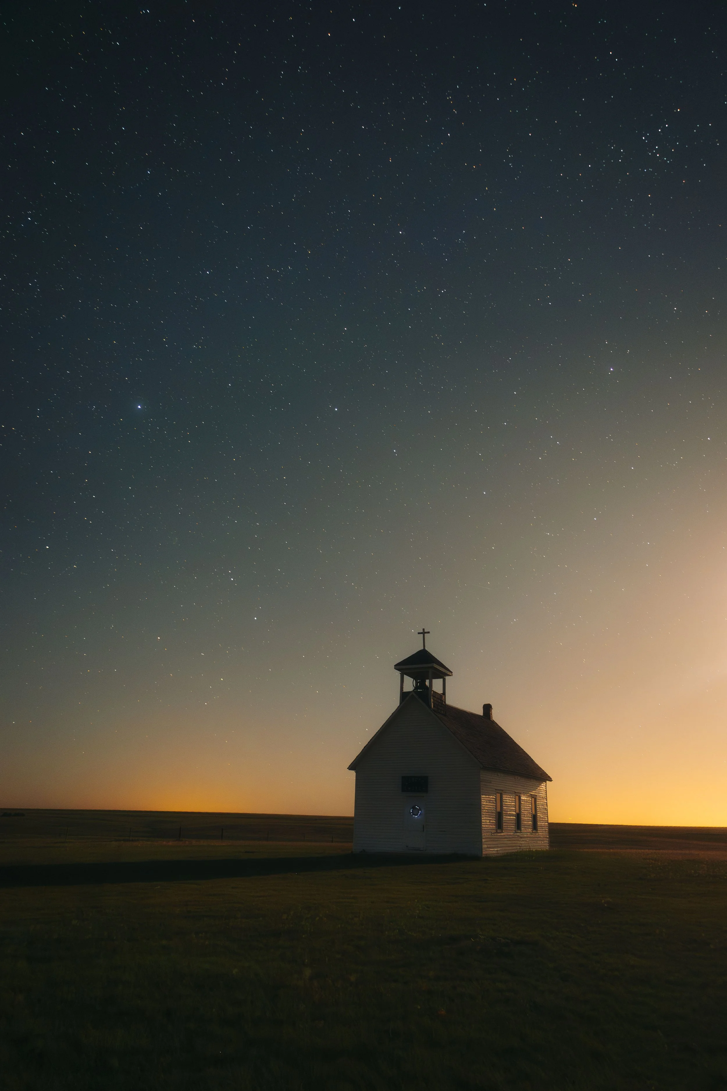 A small white church with a steeple and cross on top stands in a flat grassy field under a starry night sky with many stars visible.