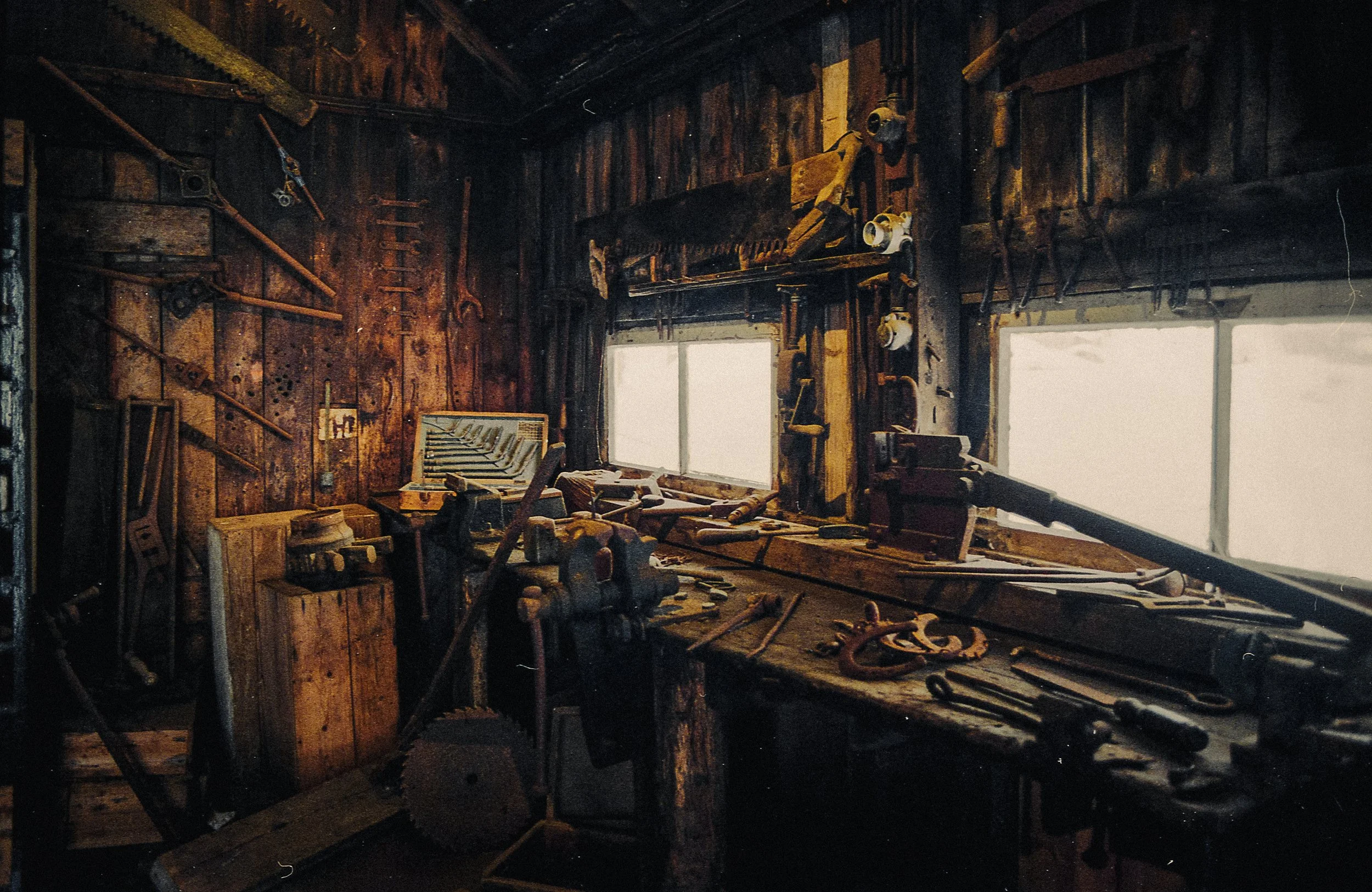 A woodworking workshop with various tools and equipment on a wooden workbench and pegboard, illuminated by natural light from two windows.