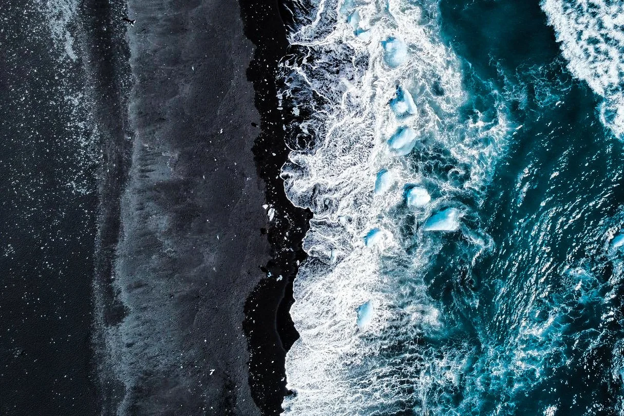 Aerial view of a black sandy beach with waves crashing onto the shore and icebergs floating in the ocean.