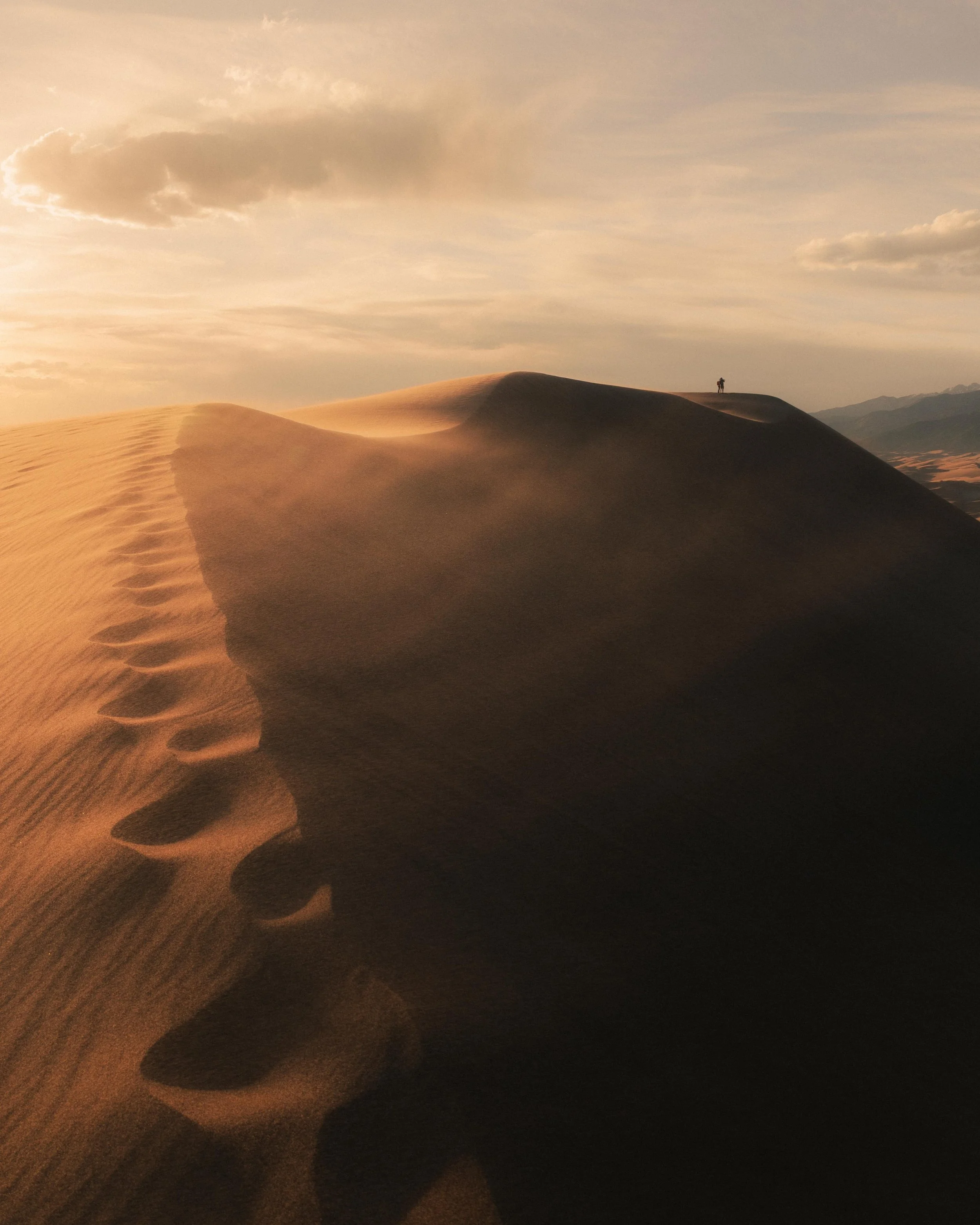 A desert landscape with sand dunes, footprints leading up a dune, a person standing at the top, under a cloudy sky during sunset.